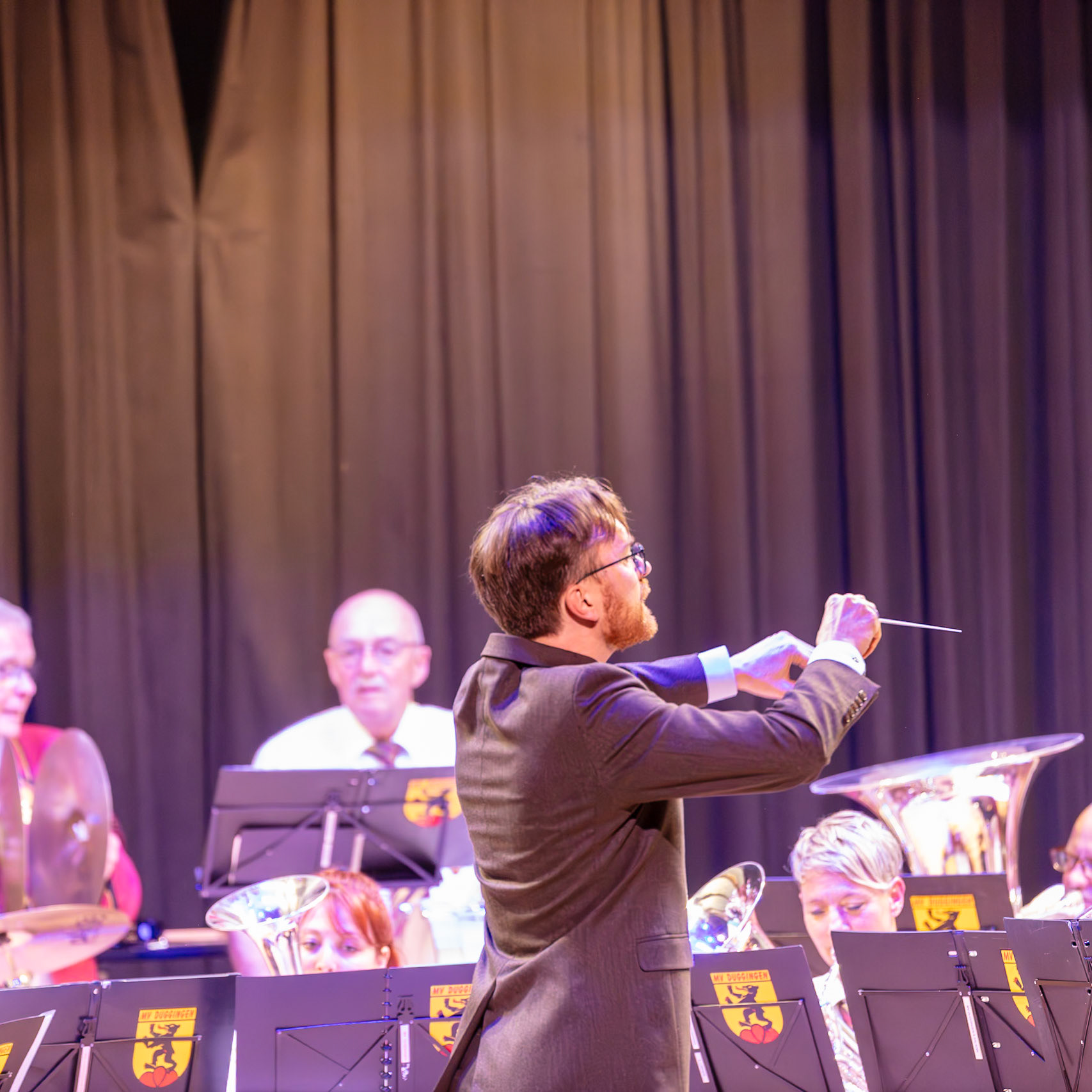 Ein Dirigent leitet ein Blasorchester.  Herr [Name des Dirigenten, falls bekannt], der Dirigent, dirigiert mit einem Dirigentenstab die Musiker des Blasorchesters.  Die Musiker spielen auf ihren Instrumenten, um Musik zu erzeugen.  Der Dirigent gibt die Musik vor und koordiniert die Musiker, damit sie synchron spielen.  Die Musik wird in einem Veranstaltungsort aufgeführt.