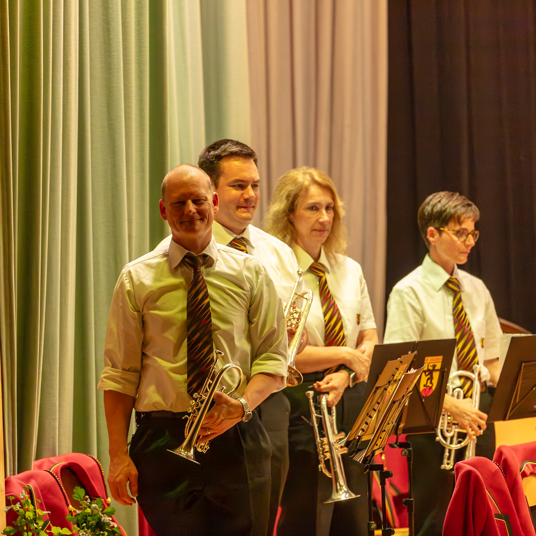 Eine Blaskapelle spielt auf der Bühne.  Herr [Name des Mannes, falls bekannt],  und die Damen [Name der Frau, falls bekannt],  sowie eine weitere Musikerin (unbekannt) spielen auf ihren Trompeten.  Sie sind in Uniform gekleidet und spielen vermutlich ein Stück Musik.  Die Atmosphäre ist konzentriert und professionell.