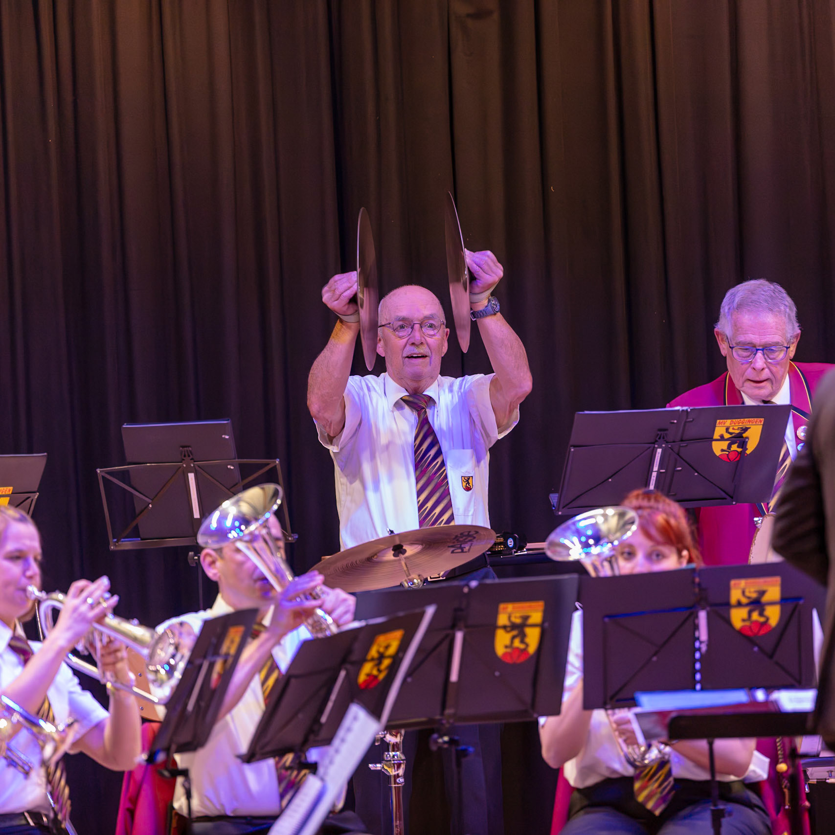 Ein Blasorchester spielt auf der Bühne.  Herr [Name des Dirigenten, falls bekannt], der Dirigent, schlägt mit zwei Becken die Musik vor.  Die Musikerinnen und Musiker des Orchesters spielen ihre Instrumente, wie Trompeten und Posaunen, um die Musik zu begleiten.  Die Atmosphäre ist konzentriert und voller Energie.