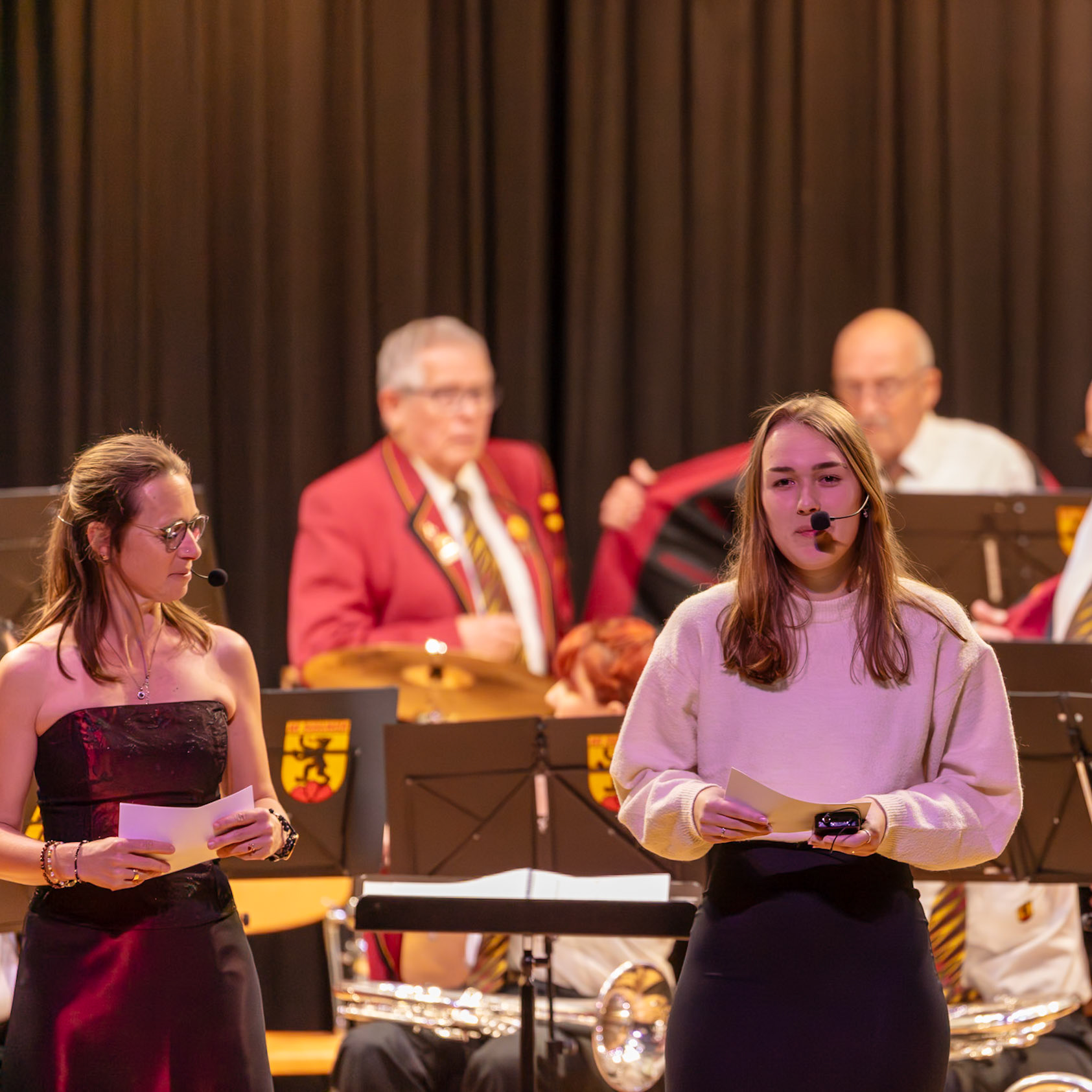 Auf der Bühne präsentieren zwei junge Frauen, vermutlich Schülerinnen oder Studentinnen, einen Auftritt.  Eine Frau in einem weinroten Kleid trägt ein Mikrofon und hält ein Blatt Papier in den Händen.  Die andere Frau, in einem cremefarbenen Pullover und schwarzen Hose, hält ebenfalls ein Blatt Papier und scheint die Moderation zu übernehmen.  Im Hintergrund ist ein Orchester oder eine Blaskapelle zu sehen, die sich auf die Aufführung vorbereitet.  Es ist wahrscheinlich ein kulturelles oder musikalisches Ereignis, bei dem die Moderatoren die Veranstaltung begleiten.