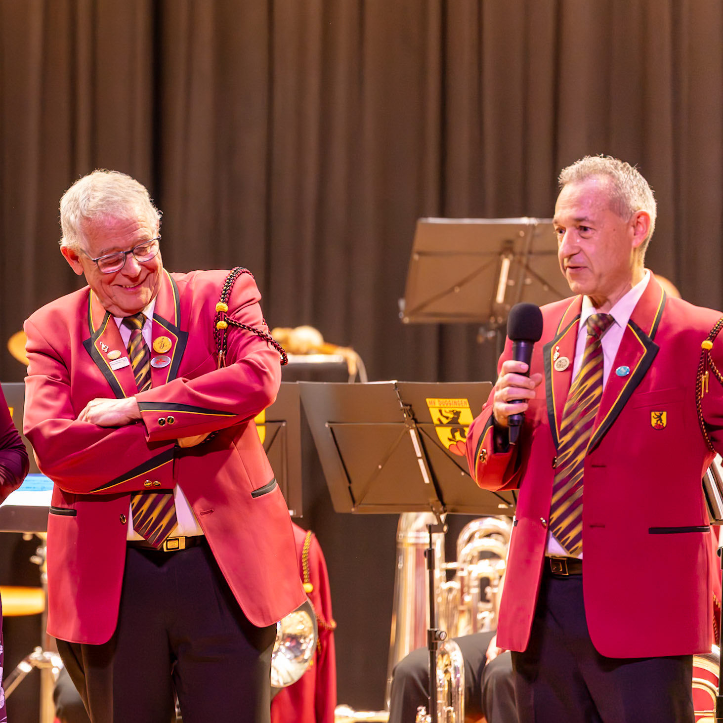 Zwei Dirigenten, Herr [Name des linken Dirigenten] und Herr [Name des rechten Dirigenten], stehen auf der Bühne vor einer Blaskapelle.  Herr [Name des rechten Dirigenten] hält ein Mikrofon und spricht vermutlich zu den Musikern.  Die Dirigenten tragen rote Uniformen, typisch für Blaskapellen, und die Musikanten sind in der Hintergrund erkennbar.  Die Szene zeigt einen Moment während eines Konzerts oder einer Übung.