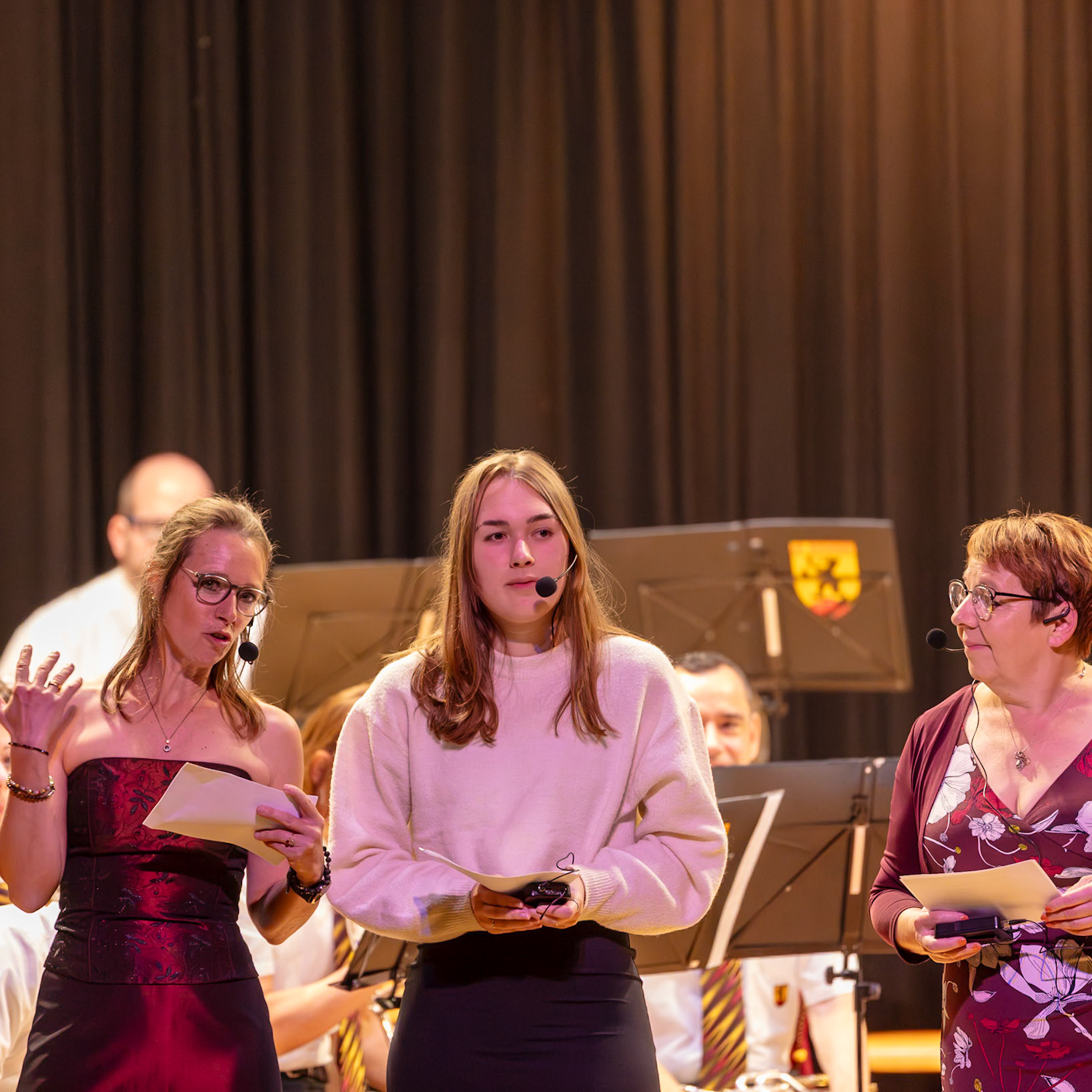 Auf der Bühne stehen drei Personen, die an einer Veranstaltung beteiligt sind.  Frau [Name der ersten Frau], vermutlich eine Moderatorin oder Rednerin, hält ein Manuskript und spricht in den Mikrofon.  Neben ihr steht die junge Frau [Name der zweiten Frau], die ebenfalls ein Mikrofon trägt und möglicherweise ebenfalls eine Rede hält oder eine musikalische Darbietung begleitet.  Frau [Name der dritten Frau] hält ebenfalls ein Manuskript und ein Mikrofon und scheint ebenfalls an der Veranstaltung beteiligt zu sein.  Wahrscheinlich handelt es sich um eine musikalische oder kulturelle Veranstaltung, bei der die Personen die Moderation oder die Begleitung der Darbietung übernehmen.