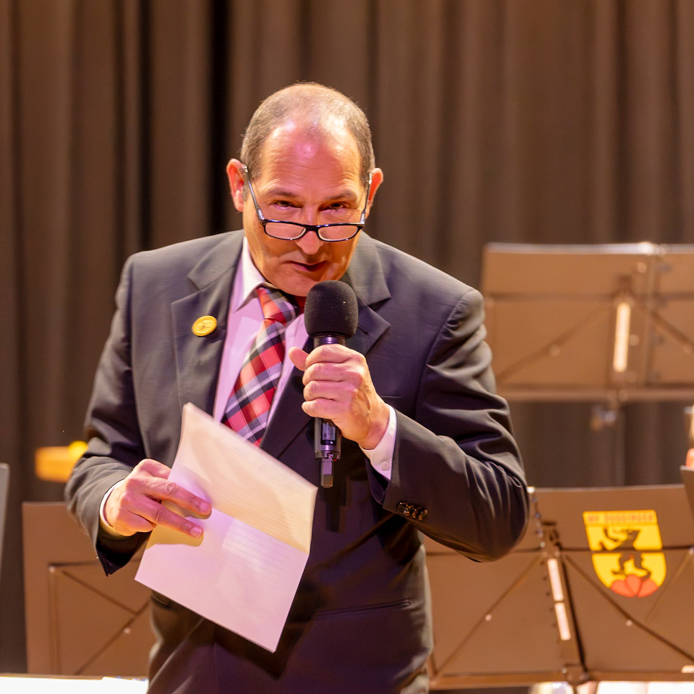 Ein Mann in Anzug, vermutlich ein Redner, hält eine Rede. Er hält ein Mikrofon und liest dabei von einem Blatt Papier.  Es ist wahrscheinlich ein Teil einer offiziellen Veranstaltung, wie z.B. einer Preisverleihung oder einer Versammlung.  Der Hintergrund deutet auf eine musikalische oder kulturelle Veranstaltung hin.