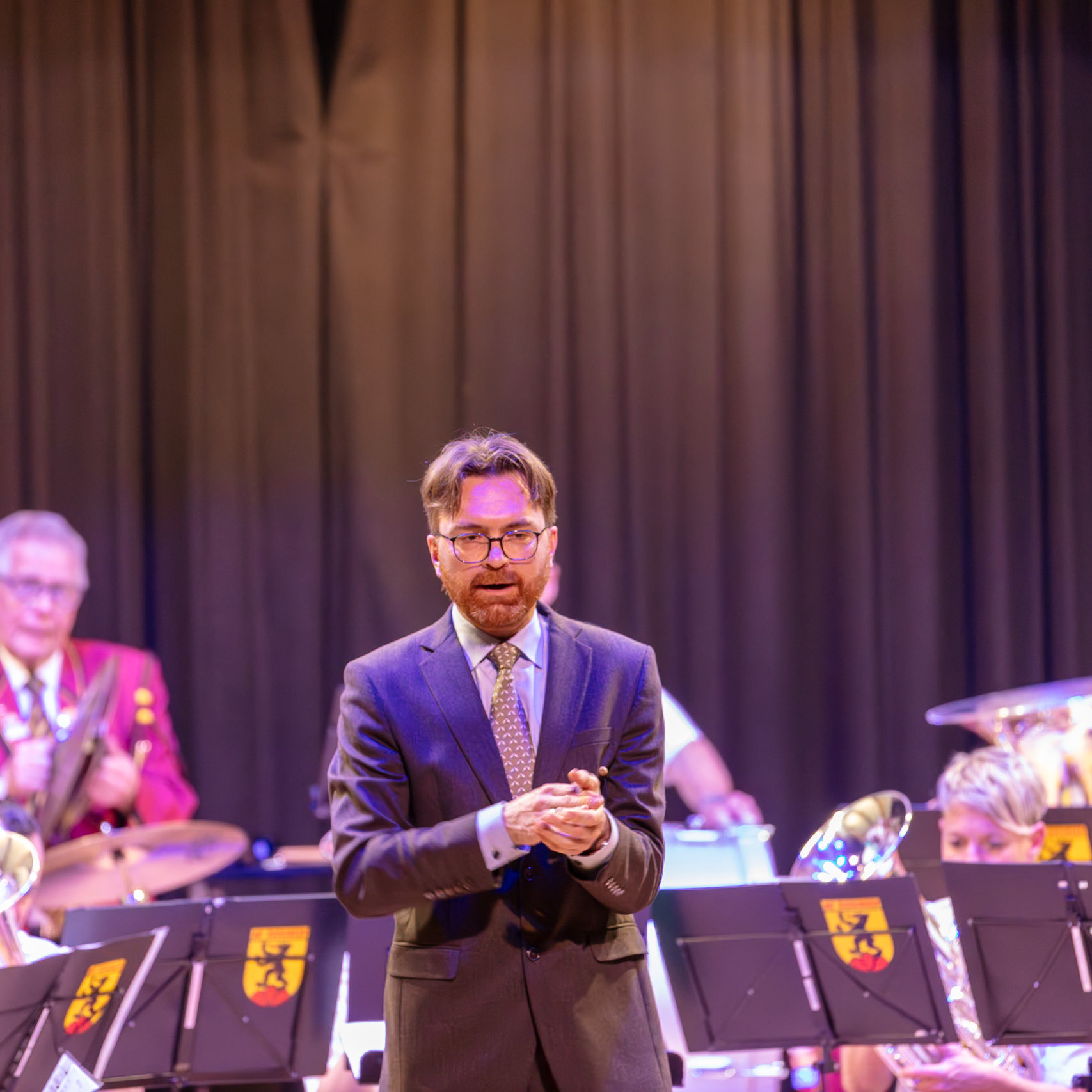 Ein Dirigent, vermutlich Herr [Name des Dirigenten], steht auf der Bühne und dirigiert ein Blasorchester.  Die Musikerinnen und Musiker des Orchesters spielen auf ihren Instrumenten.  Der Dirigent gibt Anweisungen, um die Musik zu leiten und die Harmonie des Orchesters zu gewährleisten.  Die Atmosphäre ist konzentriert und professionell, da die Musiker sich auf die Musik konzentrieren.
