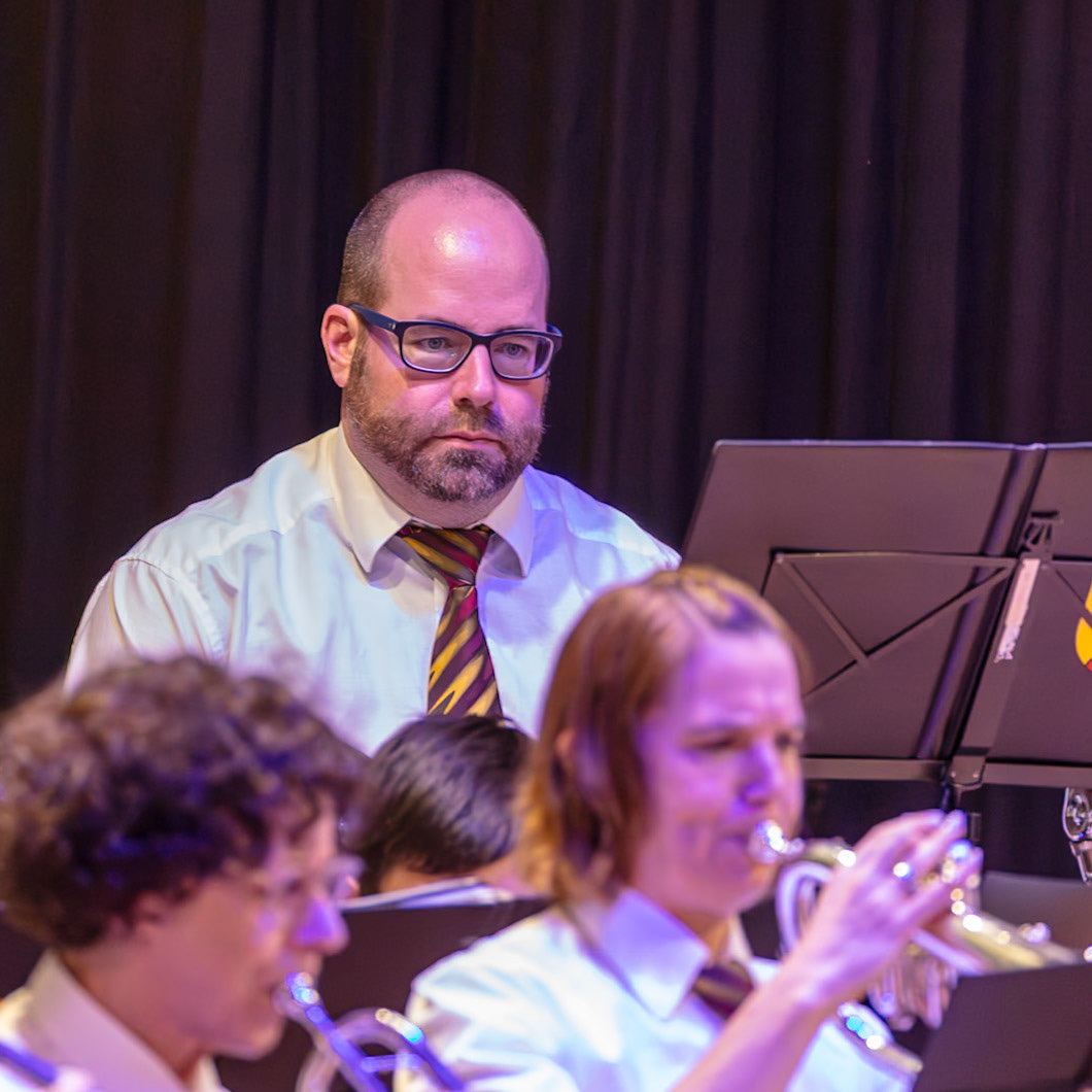 Auf der Bühne dirigiert Herr Schmidt eine Blaskapelle.  Er trägt eine weiße Bluse und einen rot-goldenen Krawatten.  Die Musikerinnen und Musiker spielen konzentriert ihre Instrumente, um die Musik zu erzeugen.  Die Atmosphäre ist professionell und voller Energie.