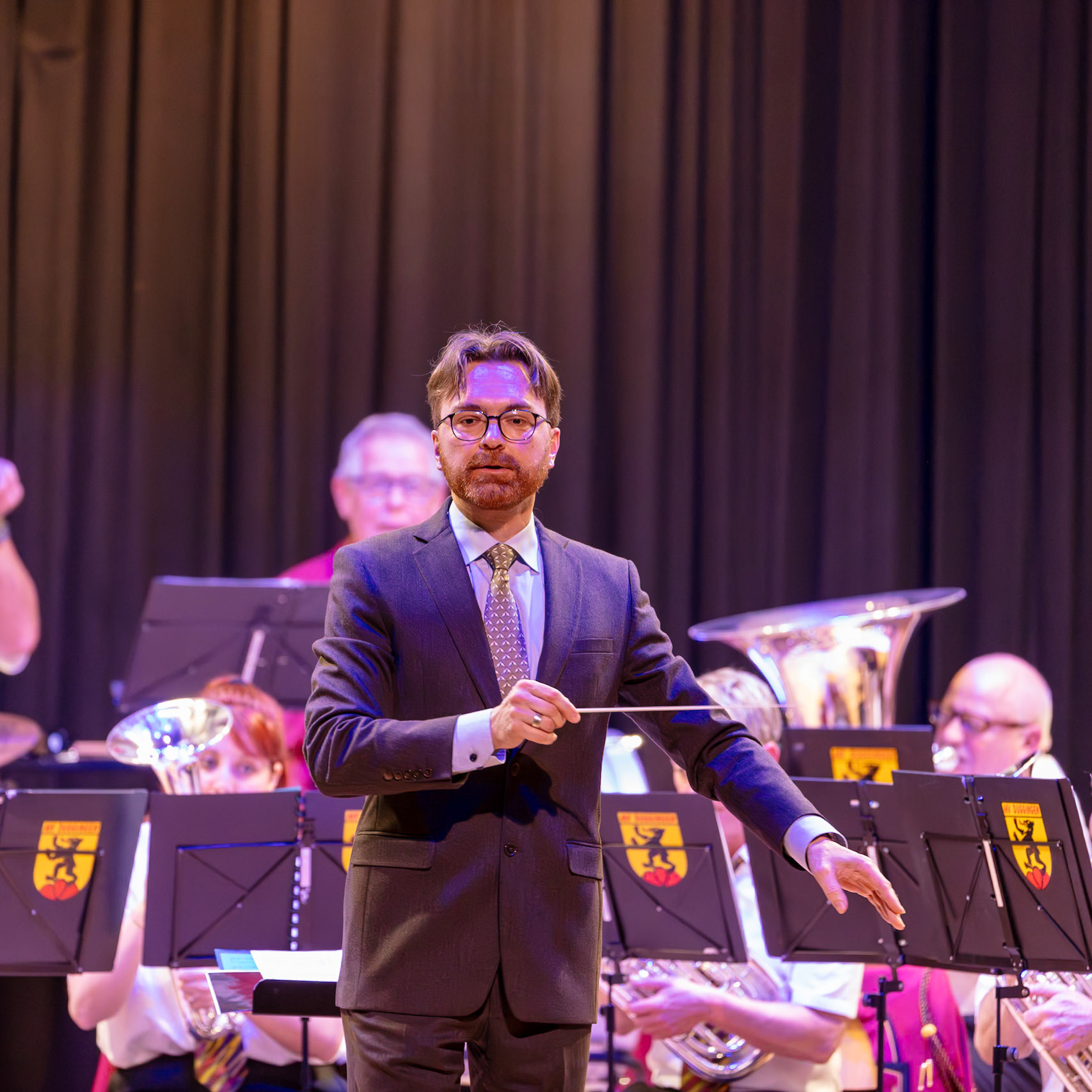 Ein Dirigent leitet ein Blasorchester. Herr [Name des Dirigenten], der Dirigent, steht auf der Bühne und dirigiert die Musiker mit seinem Dirigentenstab.  Das Blasorchester spielt Musik.  Der Dirigent gibt den Takt vor, um die Musik der Musiker zu koordinieren.  Die Musiker konzentrieren sich auf die Musik und spielen ihre Instrumente.  Die Atmosphäre ist konzentriert und professionell.