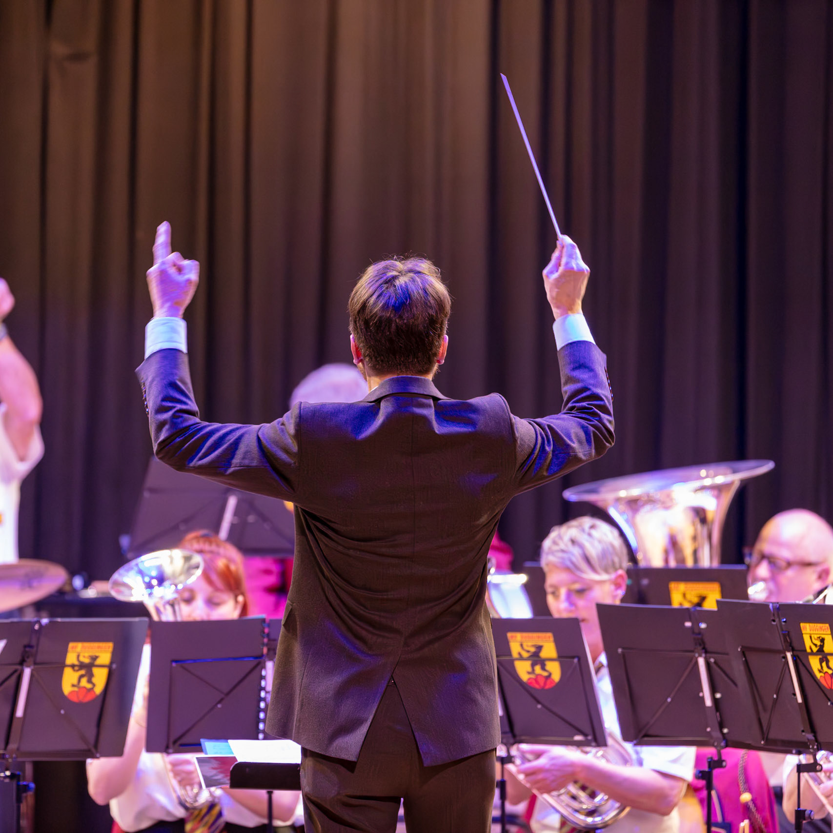 Ein Dirigent, vermutlich Herr [Name des Dirigenten, falls bekannt], leitet ein Blasorchester.  Er steht auf der Bühne und dirigiert mit einem Dirigentenstab die Musiker, die Noten vor sich auf Notenständern haben.  Die Musiker spielen verschiedene Blechblasinstrumente wie Trompeten und Posaunen.  Die Musik wird in einem Konzertsaal oder einer ähnlichen Veranstaltung aufgeführt.