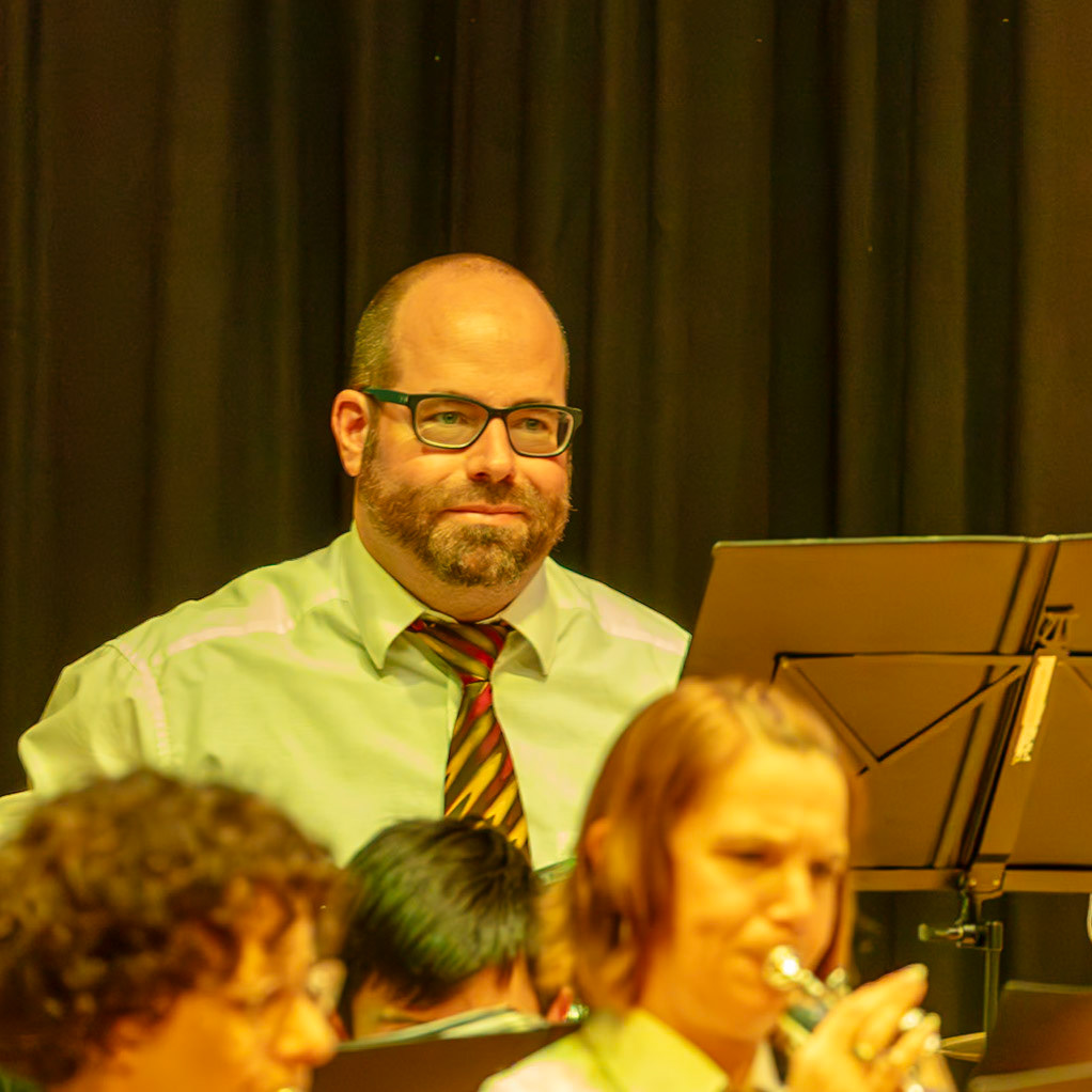 Herr Schmidt, Dirigent des Blasorchesters, leitet die Probe.  Er steht vor den Musikern und gibt Anweisungen.  Die Musiker, darunter eine Trompeterin, üben konzentriert.  Die Atmosphäre ist professionell und konzentriert, während die Musikprobe vorbereitet wird.