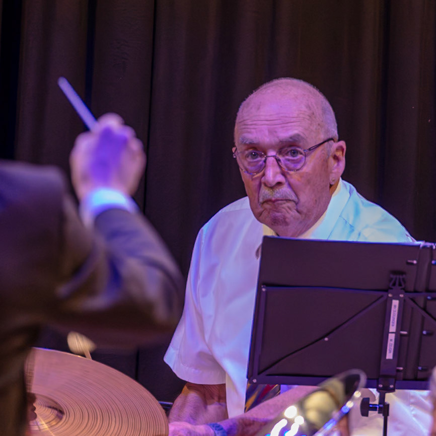 Ein Dirigent, Herr [Name des Dirigenten, falls bekannt], leitet ein Blasorchester.  Er steht vor dem Orchester und dirigiert mit einem Dirigentenstab.  Im Hintergrund ist ein Schlagzeuger zu sehen, der auf einem Schlagzeug spielt.  Die Musiker spielen Musik, und der Dirigent gibt ihnen Anweisungen, um die Musik synchron zu halten.