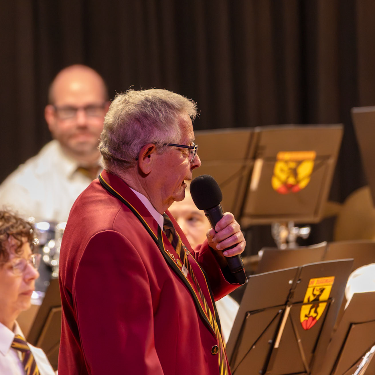 Ein Dirigent, vermutlich Herr [Name des Dirigenten, falls bekannt], spricht in die Mikrofon inmitten eines Blasorchesters.  Er trägt eine rote Uniform, typisch für Blasmusikkapellen.  Der Dirigent gibt vermutlich Anweisungen oder Ansagen an die Musiker, um die Musikaufführung zu leiten.  Die Atmosphäre ist konzentriert und professionell.