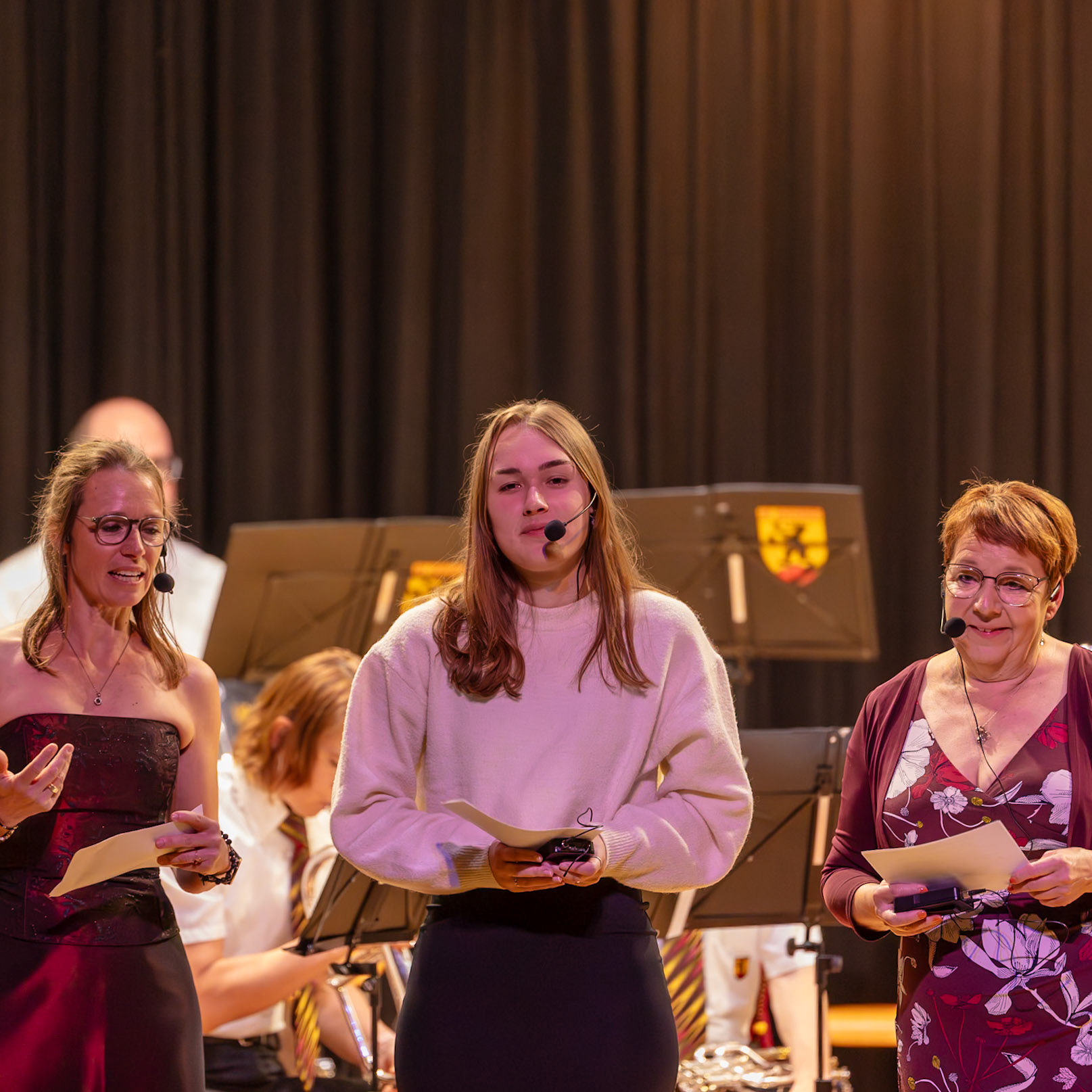 Auf der Bühne stehen drei Personen.  Frau [Name der Frau links], vermutlich Lehrerin oder Moderatorin, trägt ein rotes Kleid und hält ein Blatt Papier in der Hand.  Sie spricht in den Mikrofon und scheint eine Ansprache oder eine Zeremonie zu leiten.  Daneben steht Frau [Name der Frau rechts], ebenfalls mit einem Mikrofon, und hält ebenfalls ein Blatt Papier.  Sie scheint ebenfalls an der Moderation beteiligt zu sein.  In der Mitte steht eine junge Frau [Name der jungen Frau], die ein weißes Sweatshirt trägt und ein Blatt Papier in den Händen hält.  Sie scheint ebenfalls an der Veranstaltung beteiligt zu sein, möglicherweise als Teilnehmerin oder Leserin.  Die Personen sind Teil einer Veranstaltung, möglicherweise einer Preisverleihung oder einer ähnlichen Feierlichkeit, die von einer Musikgruppe oder einem Orchester begleitet wird.  Die Veranstaltung findet in einem Veranstaltungssaal statt.