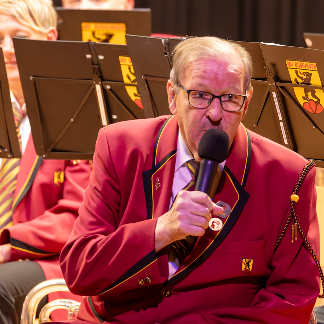 Ein Dirigent, Herr Saladín, vom Musikverein Duggingen, leitet die Musik.  Er hält ein Mikrofon und dirigiert die Musiker, die in roten Uniformen sitzen.  Die Musiker spielen vermutlich eine Blasmusik-Performance.