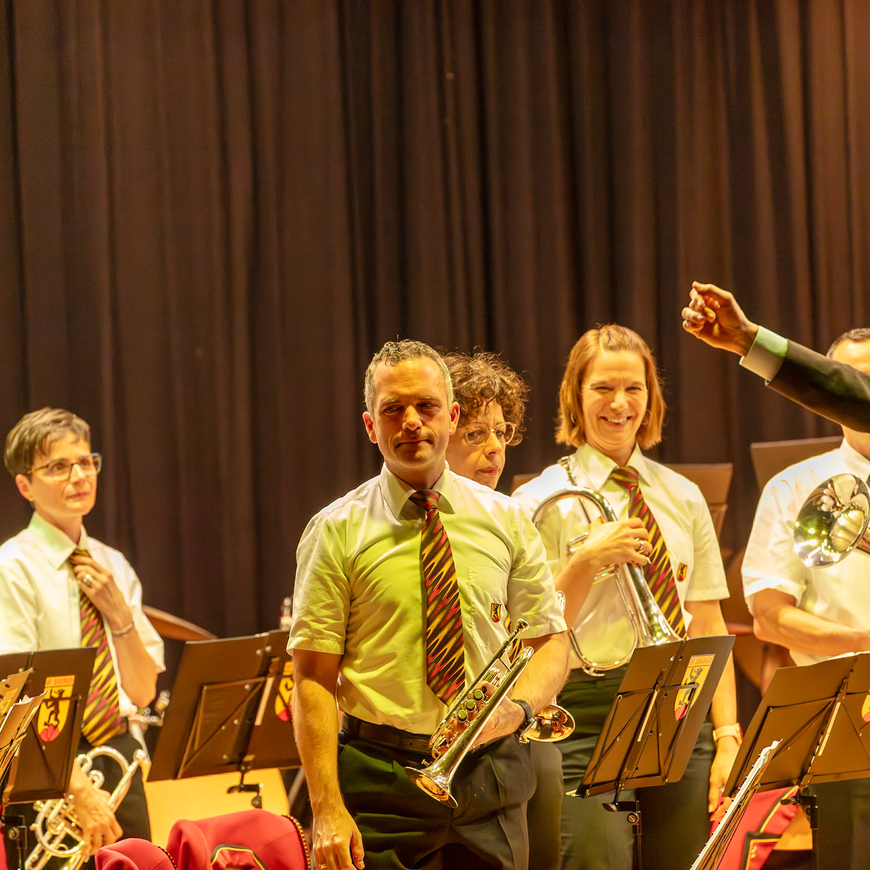 Ein Brass-Band spielt auf der Bühne.  Herr [Name des Mannes im weißen Hemd], der ein Kornett spielt, ist mittig im Bild zu sehen.  Neben ihm spielt eine Frau, vermutlich [Name der Frau], eine Trompete.  Frau [Name der Frau mit Brille], ebenfalls in der Band,  stellt ihr Krawatte zurecht.  Alle Musiker sind in einheitlichen weißen Hemden und Krawatten gekleidet, was auf eine organisierte Band hinweist.  Die Band spielt vermutlich für ein Publikum, das nicht im Bild zu sehen ist.