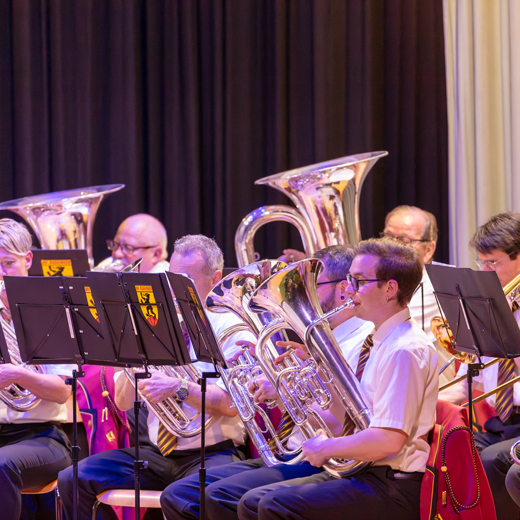 Eine Blaskapelle spielt auf der Bühne.  Musikerinnen und Musiker, vermutlich Mitglieder der [Name der Kapelle], sitzen auf Stühlen und spielen auf ihren jeweiligen Instrumenten, hauptsächlich Tuba und Euphonium.  Die Musiker tragen einheitliche, rote Jacken mit goldenen Details und schwarze Hosen.  Sie spielen, um ein Publikum zu unterhalten.