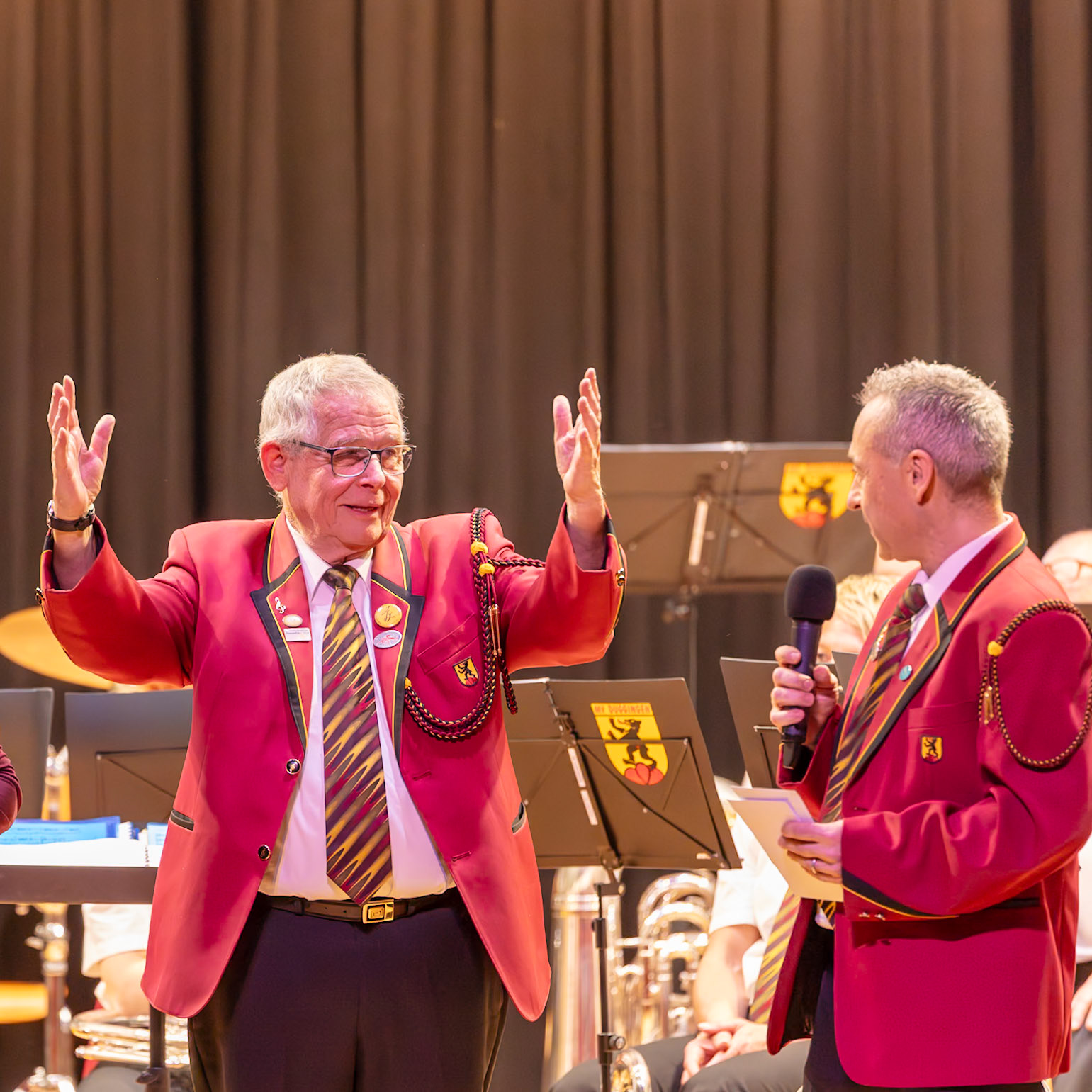Zwei Personen in roten Uniformen, vermutlich Mitglieder eines Musikvereins, stehen auf einer Bühne.  Herr [Name des Mannes links], Dirigent, dirigiert mit erhobenen Händen die Blaskapelle.  Er hält ein Mikrofon und spricht vermutlich eine Ansprache oder gibt Anweisungen an die Musiker.  Die Kapelle spielt, wie man an den Instrumenten im Hintergrund erkennen kann.  Die rote Uniform und die Embleme deuten auf eine offizielle Funktion oder Mitgliedschaft im Verein hin.  Der Hintergrund zeigt die Bühne und die Musiker.