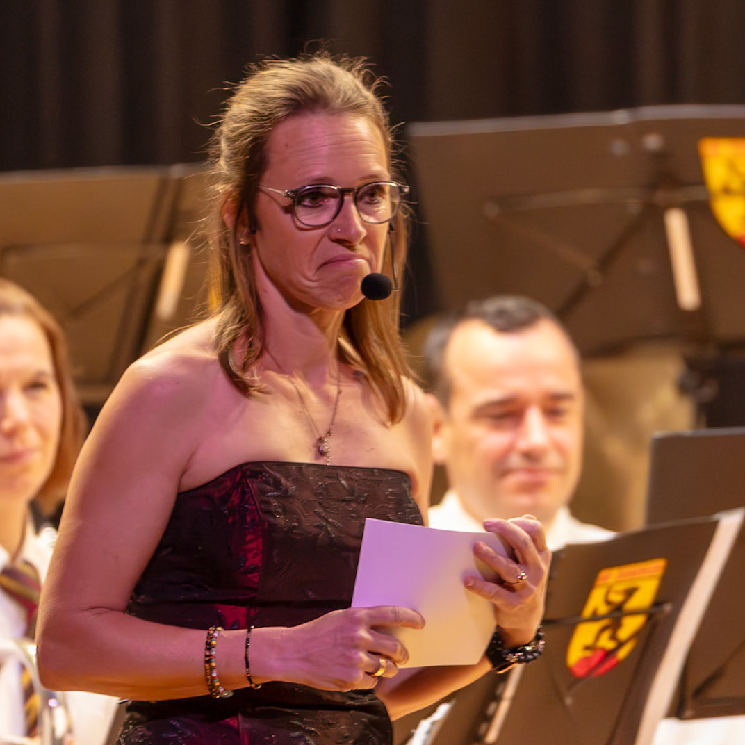 Eine Dirigentin, vermutlich Frau Schmidt, hält eine Partitur in der Hand und dirigiert eine Blaskapelle.  Sie trägt eine rote, mit Mustern verzierte Abendrobe und eine Kette.  Die Dirigentin steht vor einem Dirigentenpult und spricht in das Mikrofon.  Der Hintergrund zeigt weitere Musiker der Kapelle.  Der Moment zeigt die Konzentration und die Vorbereitung auf die musikalische Darbietung.