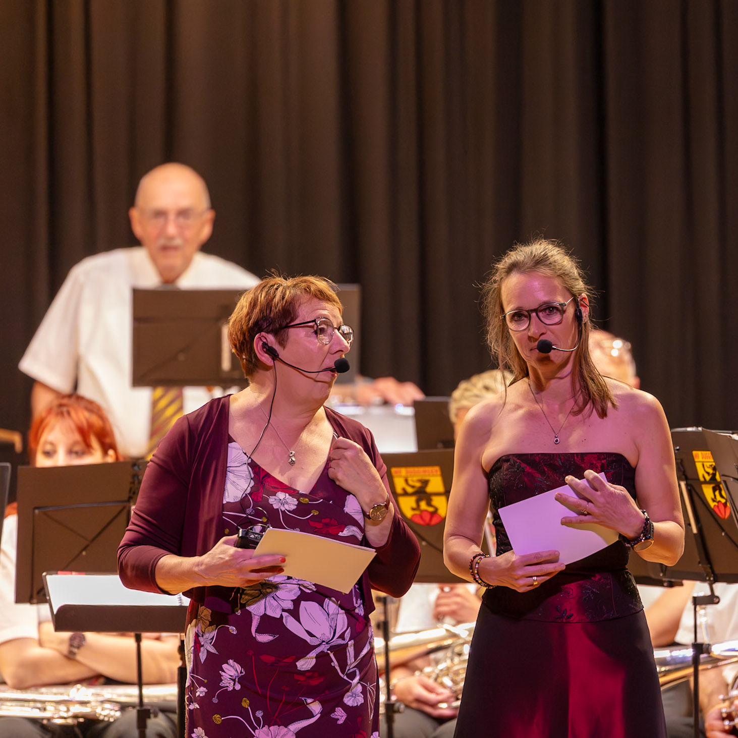 Zwei Damen, vermutlich Moderatorinnen, leiten ein Konzert.  Frau [Name der Frau links], mit ihrem Mikrofon und Noten, und Frau [Name der Frau rechts], mit einem weiteren Mikrofon und einem Blatt Papier, präsentieren oder moderieren die Veranstaltung.  Sie unterstützen die Musiker/innen des Blasorchesters, das im Hintergrund zu sehen ist.  Der Anlass ist vermutlich ein Konzert oder eine ähnliche musikalische Veranstaltung.