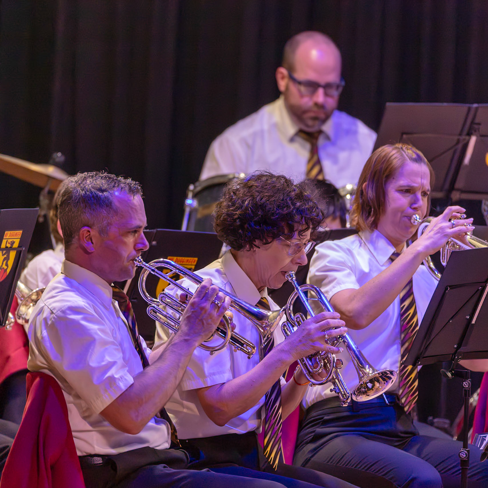 Eine Blaskapelle spielt auf der Bühne.  Mehrere Musikerinnen und Musiker, darunter [Name des Mannes links], spielen auf ihren Trompeten und Kornetten.  Sie tragen einheitliche weiße Hemden und Krawatten.  Die Musikerinnen und Musiker konzentrieren sich auf die Musik und spielen mit Leidenschaft.  Die Atmosphäre ist lebendig und voller Energie.
