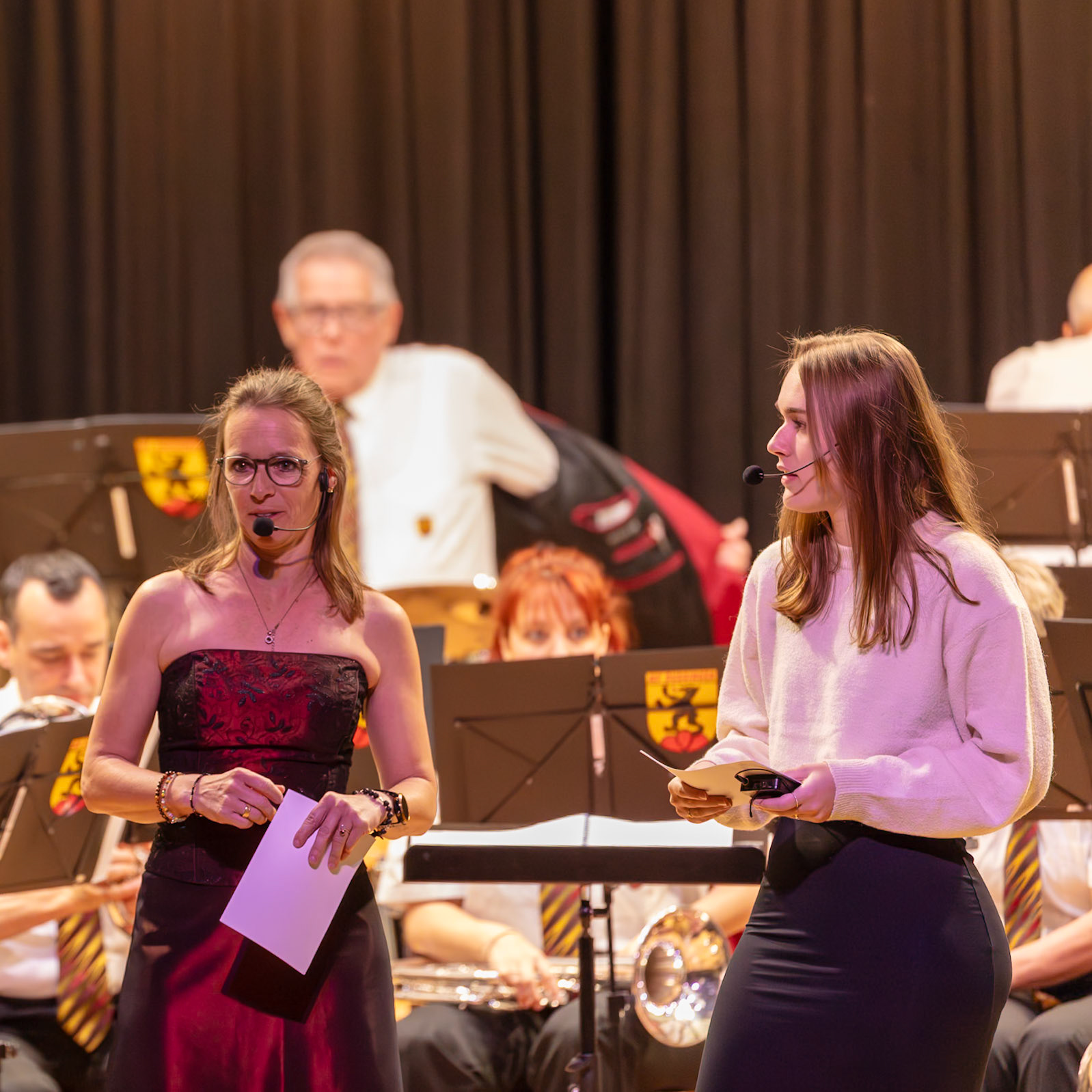 Auf der Bühne präsentieren zwei Personen, Frau [Name der Frau] (Moderatorin) und [Name des Mädchens] (möglicherweise eine weitere Moderatorin oder Sprecherin), die Leistungen eines Musikvereins. Frau [Name der Frau] hält ein Blatt Papier in den Händen, während [Name des Mädchens] in den Mikrofonen spricht.  Sie präsentieren wahrscheinlich die einzelnen Musiker oder Gruppen des Musikvereins und/oder würdigen deren Leistungen.  Die Veranstaltung findet vor einem Publikum statt, das durch die unscharfen Hintergründe der Musiker erkennbar ist.  Der Grund für die Präsentation ist die Anerkennung und Würdigung der musikalischen Darbietungen des Musikvereins.