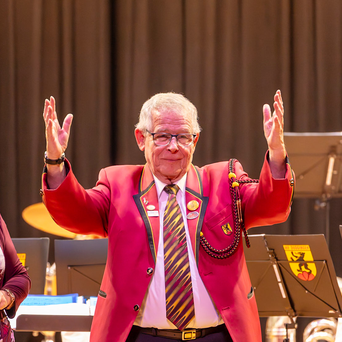 Herr [Name des Dirigenten], der Dirigent des Blasorchesters, dirigiert mit erhobenen Händen.  Er trägt ein rotes, festliches Jacket und einen auffälligen Krawattenmuster.  Die Geste deutet auf eine enthusiastische und engagierte musikalische Leistung hin, die wahrscheinlich gerade stattfindet oder kurz bevorsteht.  Die Umgebung deutet auf ein Konzert oder eine ähnliche Veranstaltung in einem Veranstaltungssaal hin.