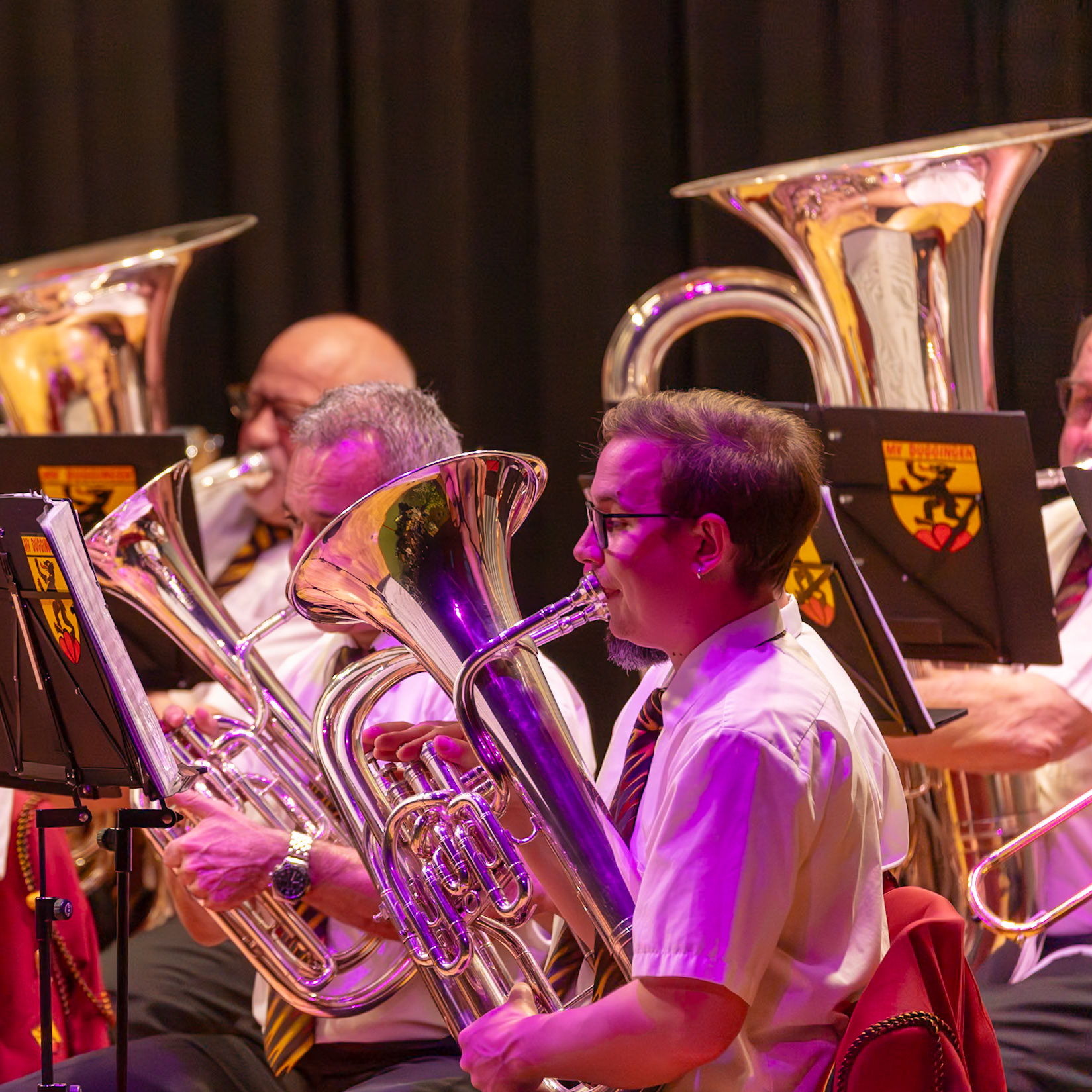 Auf der Bühne spielt ein Blasorchester.  Hier spielen mehrere Musiker, darunter [Name des Musikers], der ein Tenorhorn spielt.  Die Musiker konzentrieren sich auf die Ausführung der Musik, die von den Noten auf ihren Notenständern bestimmt wird.  Die Atmosphäre ist lebendig und konzentriert, während die Musik erklingt.