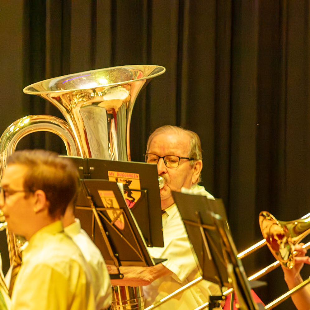 Eine Blaskapelle spielt auf.  Herr [Name des Musikers, falls bekannt], der Tuba spielt, konzentriert sich auf die Musik.  Andere Musiker, darunter [Name des Musikers, falls bekannt], spielen ebenfalls ihre Instrumente.  Die Musiker der Blaskapelle, wahrscheinlich Teil des Musikvereins "My Duggingen", üben oder spielen ein Konzert in einem Veranstaltungsort.