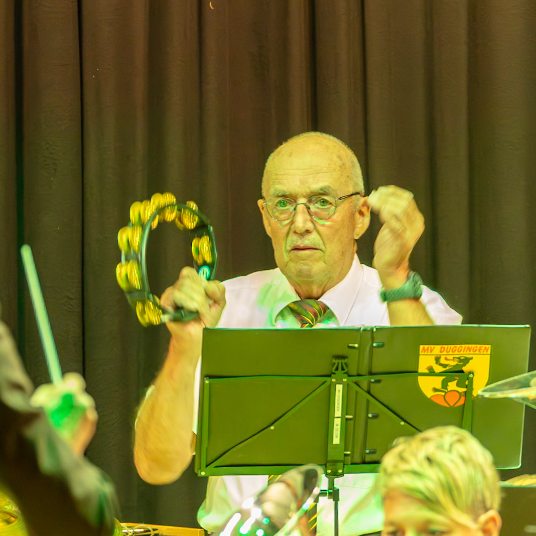 Ein Dirigent der Musikvereinigung Duggingen leitet die Gruppe.  Er trägt eine weiße Bluse und einen Krawatte mit grünen und schwarzen Streifen.  Er hält eine Tamburin in der Hand und gibt Anweisungen an die Musiker.  Die Musikvereinigung Duggingen ist erkennbar an ihrem Emblem auf dem Notenständer.