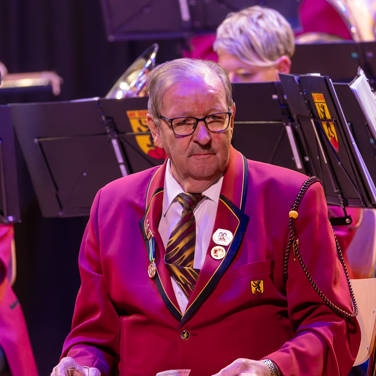 Herr [Name des Mannes], Dirigent des Blasorchesters, ist inmitten der Musiker*innen zu sehen. Er trägt eine rote Uniform mit goldenen Details und Ehrenzeichen.  Offensichtlich leitet er das Orchester, denn er ist konzentriert und blickt auf die Musiker*innen.  Die Uniform und die Anwesenheit des Dirigenten deuten auf ein Konzert oder eine Übung eines Blasorchesters hin.  Die sichtbaren Abzeichen deuten auf eine Jubiläumsfeier oder eine besondere Anerkennung hin.  Die Atmosphäre ist professionell und konzentriert.