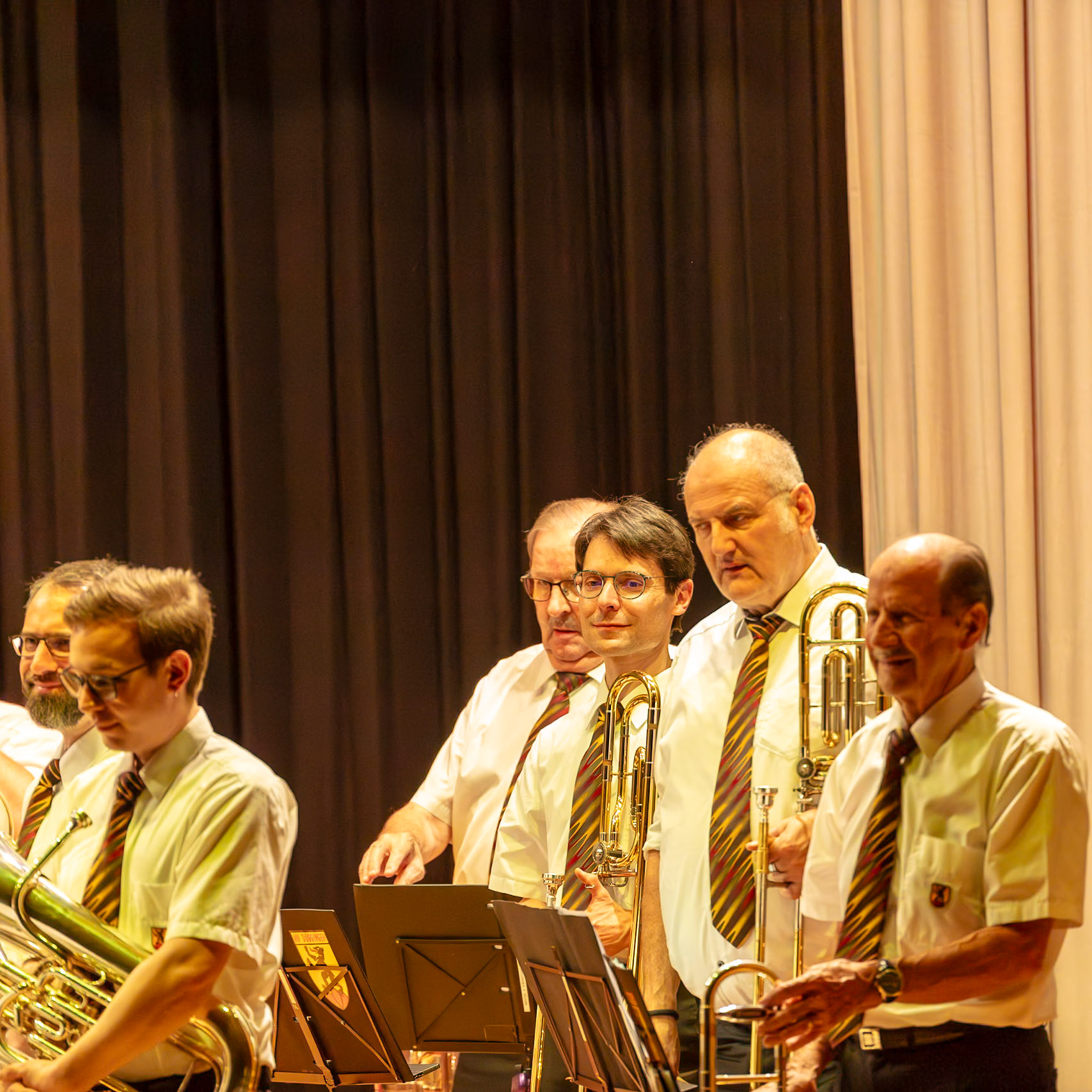 Auf der Bühne einer Veranstaltung spielen mehrere Musiker eines Blasorchesters.  Herr Meier (Trompete), Herr Schmidt (Posaune) und Herr Müller (Tenorhorn) sind konzentriert dabei, Musik zu machen.  Sie tragen einheitliche, helle Hemden und farbige Krawatten.  Die Atmosphäre ist professionell und konzentriert, während die Musiker ihre Instrumente spielen.  Es ist ein gemeinsames Musizieren, das Freude und Harmonie ausstrahlt.