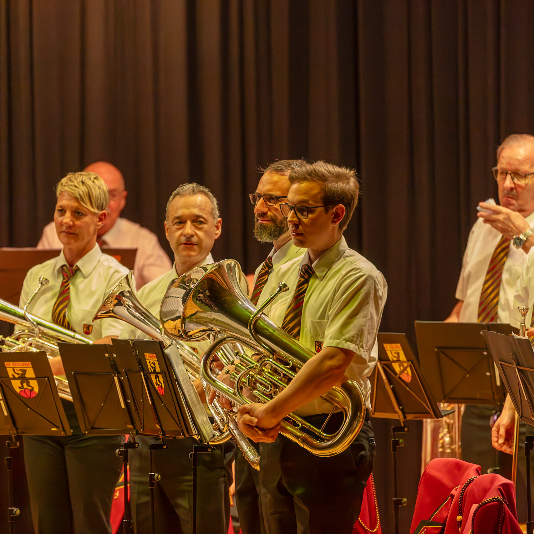 Ein Blasorchester spielt auf der Bühne.  Mehrere Musiker, darunter  (Name des Musikers 1), (Name des Musikers 2) und (Name des Musikers 3),  spielen auf ihren jeweiligen Blechblasinstrumenten, wie z.B.  Euphonien.  Sie tragen einheitliche, weiße Hemden und farbige Krawatten.  Die Musiker konzentrieren sich auf die Ausführung der Musik, die von den Noten auf ihren Notenständern bestimmt wird.  Die Atmosphäre ist professionell und konzentriert.