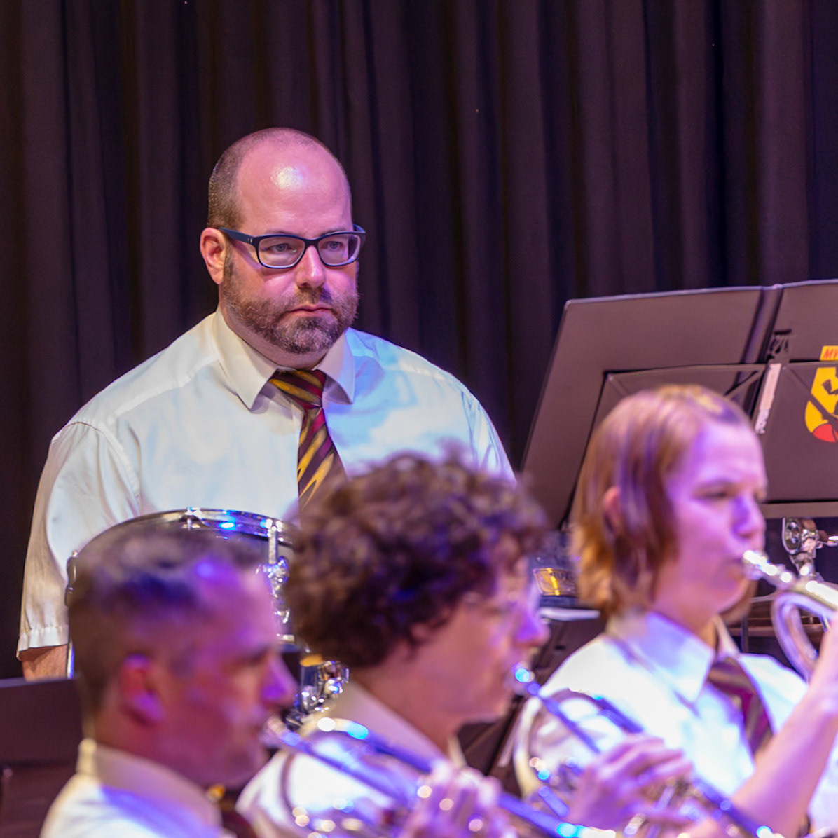Ein Blasorchester spielt auf der Bühne. Herr [Name des Dirigenten/Leiters], erkennbar an seiner Brille und dem Krawattenmuster, dirigiert die Musiker.  Die Musikerinnen und Musiker spielen auf ihren Instrumenten, darunter Trompeten und wahrscheinlich weitere Blechblasinstrumente.  Die Musik wird von den Zuschauern genossen.  Die Atmosphäre ist konzentriert und voller musikalischer Energie.