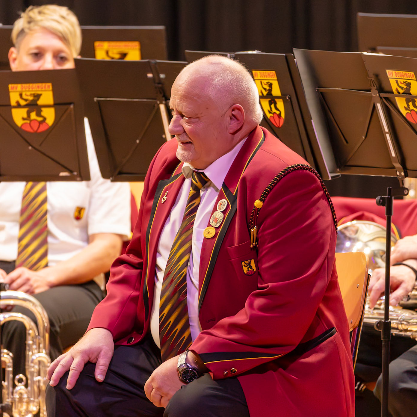 Ein Dirigent, vermutlich Herr [Name des Dirigenten, falls bekannt], sitzt inmitten eines Blasorchesters und dirigiert.  Er trägt ein rotes, festliches Jacket mit goldenen Akzenten und einem auffälligen, gemusterten Krawatten.  Die Musiker des Orchesters sind in ihren Uniformjacken und sitzen an ihren Instrumenten.  Die Atmosphäre ist konzentriert und professionell, da sich alle auf die Musik konzentrieren.  Der Dirigent gibt Anweisungen und leitet das Orchester, um die Musik zu spielen.