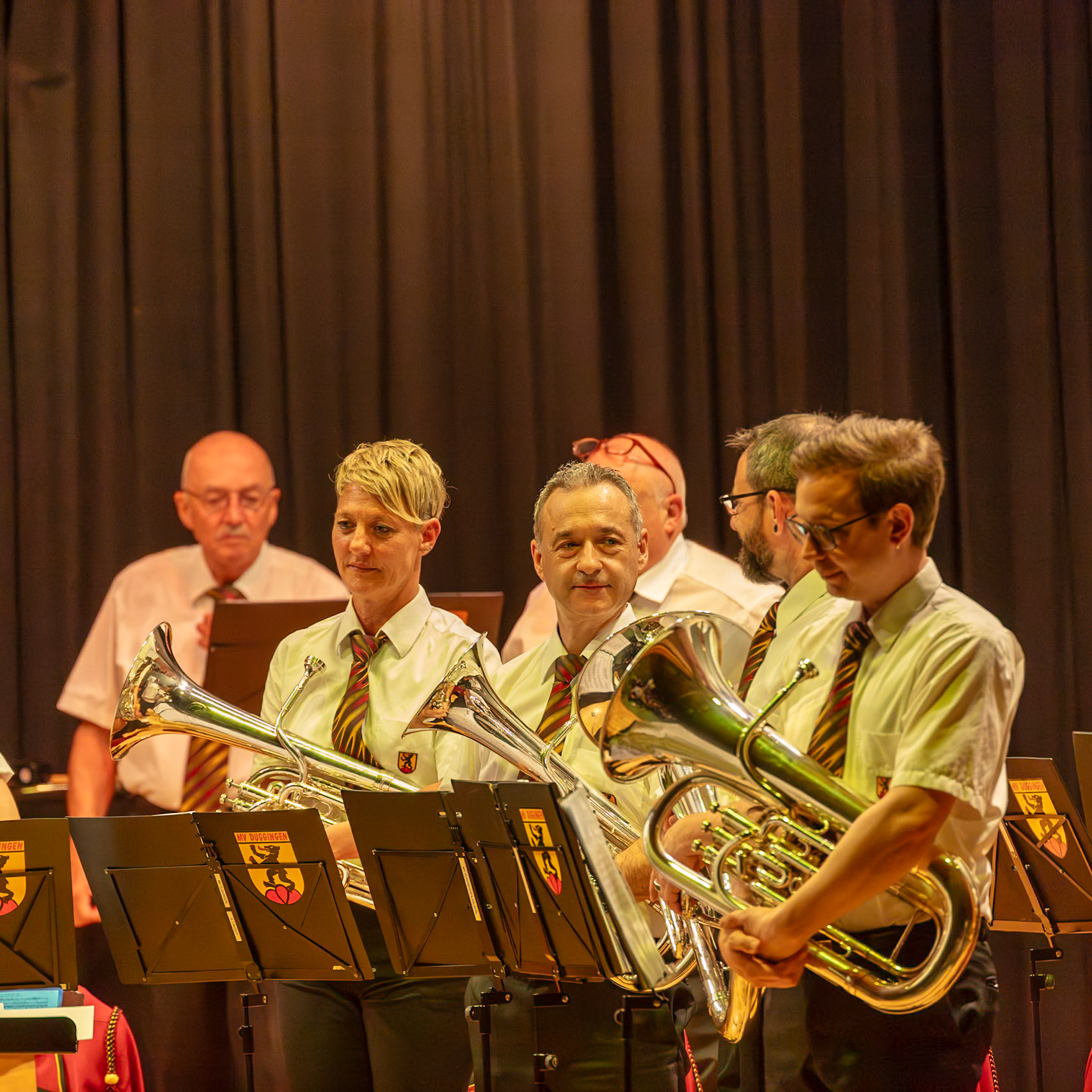 Eine Blaskapelle spielt auf der Bühne.  Die Musiker, vermutlich Mitglieder der MV Duggingen, tragen einheitliche weiße Hemden und farbige Krawatten.  Sie spielen auf ihren jeweiligen Instrumenten, hier insbesondere auf Tuba und Flügelhörnern.  Die Musik wird vermutlich für ein Publikum aufgeführt, das in der Halle sitzt.  Die Konzentration der Musiker auf ihre Instrumente und die Stimmung im Raum deuten auf eine ernsthafte und konzentrierte Aufführung hin.