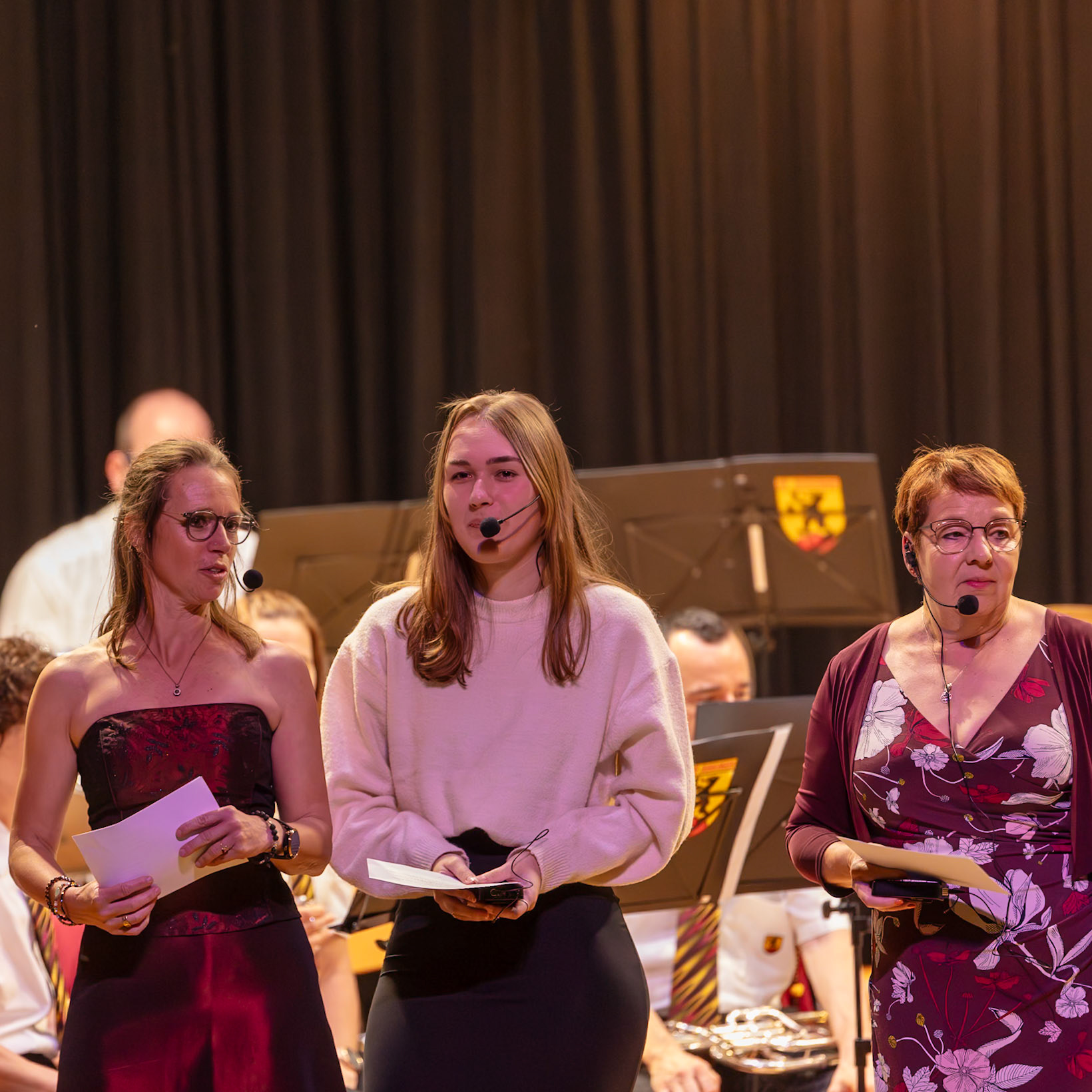 Drei Personen sind bei einer musikalischen Veranstaltung aktiv.  Frau [Name der Frau links], vermutlich eine Lehrerin oder Verantwortliche, hält ein Blatt Papier und spricht in den Mikrofon.  Frau [Name der Frau Mitte], ebenfalls mit Mikrofon, scheint die Veranstaltung zu leiten oder zu moderieren.  Frau [Name der Frau rechts], ebenfalls mit Mikrofon, ist möglicherweise Teil des Organisationsteams oder einer Sprecherin.  Sie alle tragen Kleidung, die für eine formelle Veranstaltung geeignet ist.  Der Hintergrund deutet auf eine Schule oder eine ähnliche Institution hin.  Die Personen sind dabei, die Veranstaltung zu leiten oder zu unterstützen.
