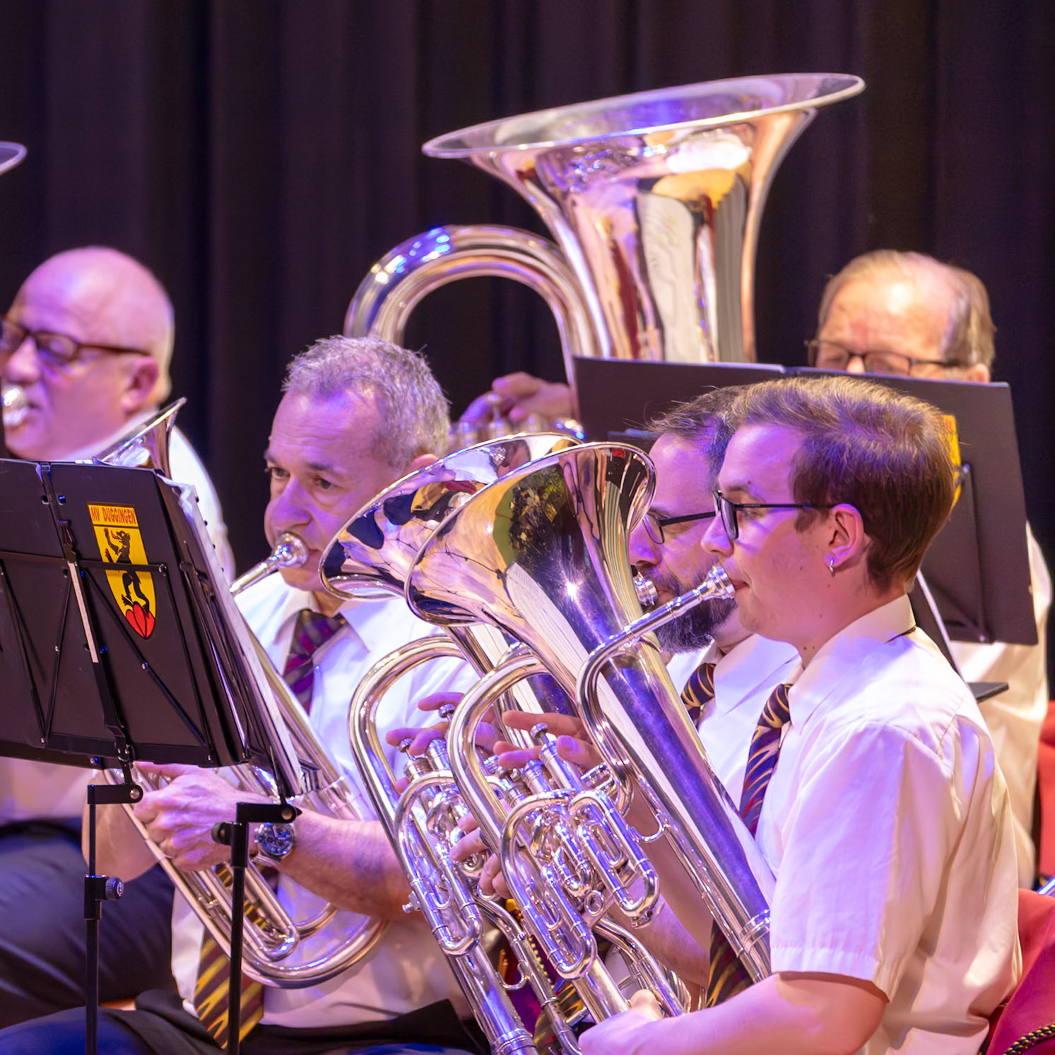 Eine Blaskapelle spielt auf.  Mehrere Musiker, darunter [Name des Musikers 1] (Trompeter), [Name des Musikers 2] (Posaunist) und [Name des Musikers 3] (Euphoniumspieler), spielen auf ihren Instrumenten.  Sie tragen einheitliche weiße Hemden und Krawatten.  Die Musiker konzentrieren sich auf die Ausführung der Musik, die von den Instrumenten erzeugt wird.  Die Atmosphäre ist konzentriert und voller musikalischer Energie.