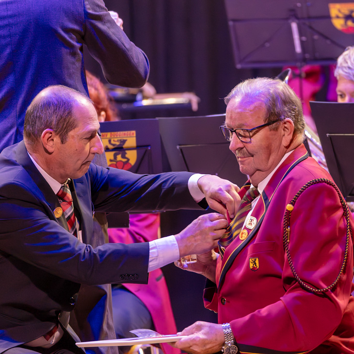 Herr [Name des Empfängers], vermutlich ein Mitglied des Musikzuges, erhält von Herrn [Name des Überreichenden] die Auszeichnung.  Die Auszeichnung wird an der Uniform befestigt.  Es ist ein feierlicher Moment, wahrscheinlich im Rahmen einer Ehrung oder einer Zeremonie für Verdienste um den Musikzug.