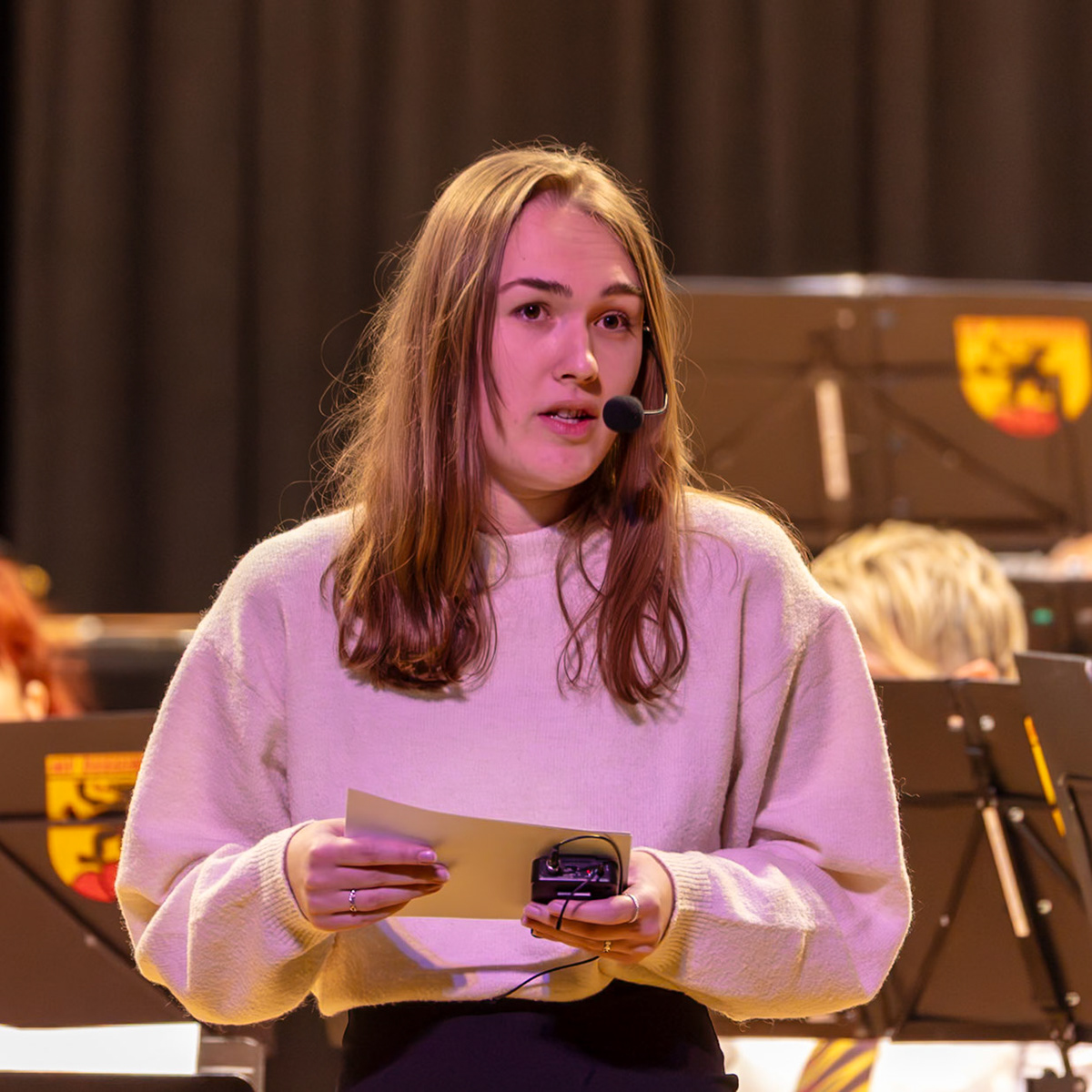 Eine junge Frau, vermutlich eine Schülerin namens [Name der Schülerin], liest eine Ansprache vor einem Orchester.  Sie trägt ein Mikrofon und hält Notizen in den Händen.  Sie leitet wahrscheinlich die musikalische Darbietung oder gibt eine Ansprache an die Musiker oder das Publikum.