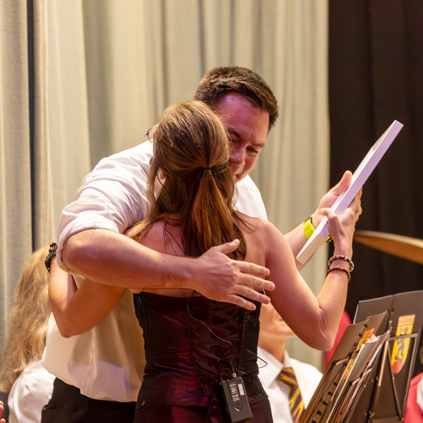 Zwei Personen, vermutlich Dirigent und Dirigentin, umarmen sich und tauschen etwas aus.  Die Dirigentin (Name unbekannt) übergibt dem Dirigenten (Name unbekannt) ein Notenblatt.  Dies geschieht vermutlich nach einer erfolgreichen Probe oder einem Auftritt, als Zeichen der Wertschätzung und des Zusammenhalts im Orchester.