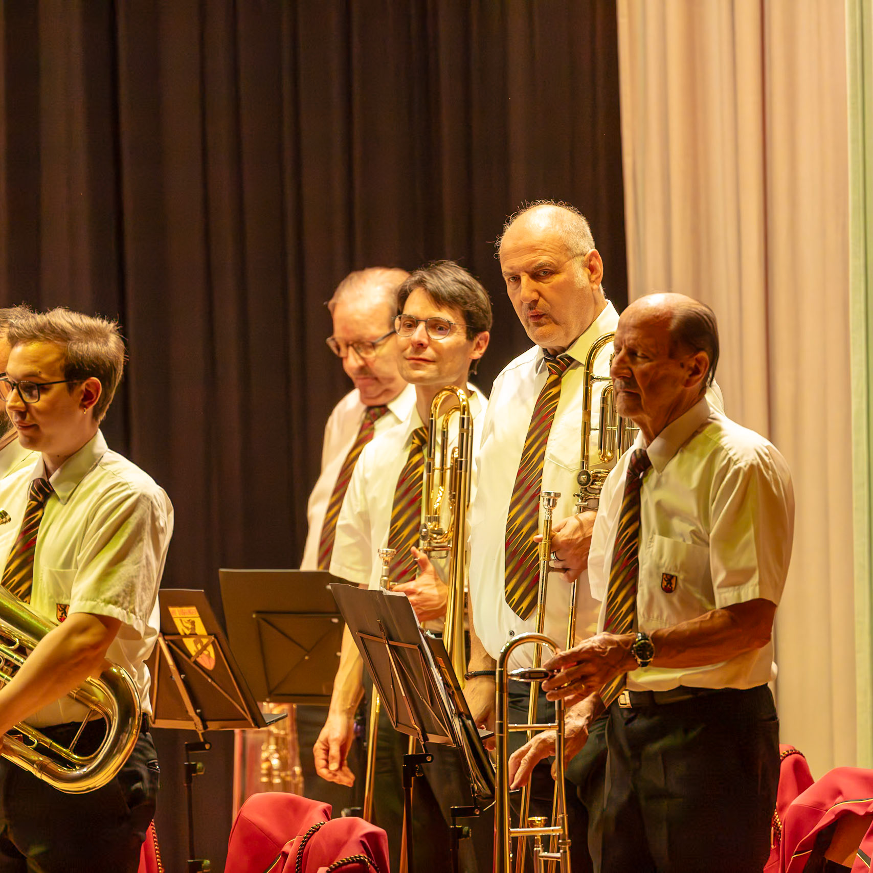 Auf der Bühne spielen mehrere Musiker eines Blasorchesters.  Die Trompeter, mit ihren goldenen Instrumenten, und der Tuba-Spieler konzentrieren sich auf die Musik.  Sie tragen einheitliche, weiße Hemden und farbige Krawatten.  Die Atmosphäre ist konzentriert und professionell, während die Musiker ihre Instrumente spielen.