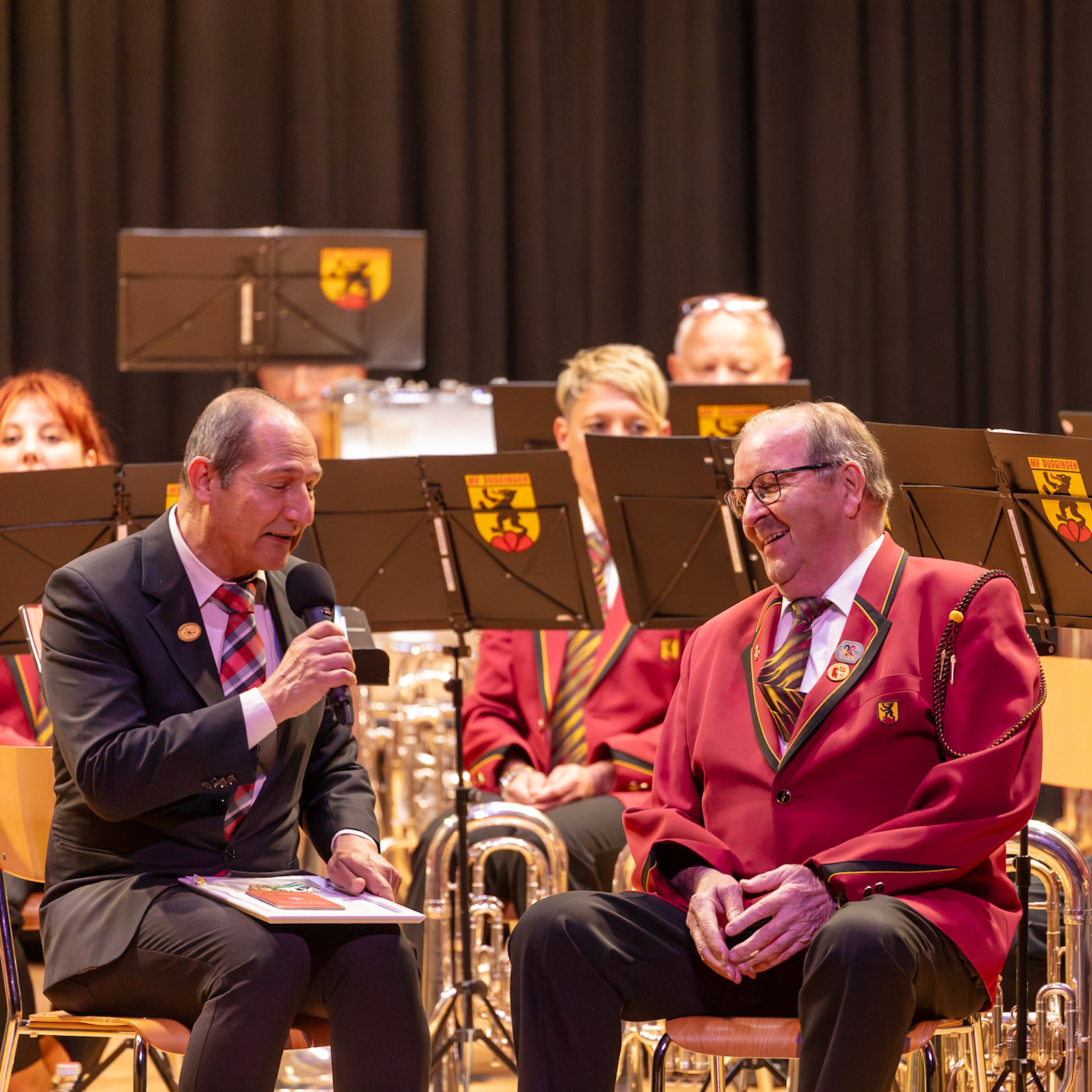Auf der Bühne findet eine Veranstaltung mit einem Blasorchester statt. Herr [Name des Mannes im dunklen Anzug], vermutlich ein Ehrengast oder Moderator, hält ein Mikrofon und spricht mit dem Publikum.  Er ist inmitten der Musiker des Blasorchesters, die in roten Uniformen sitzen.  Der Anlass ist wahrscheinlich ein Konzert oder eine ähnliche Veranstaltung, bei der Herr [Name des Mannes im dunklen Anzug] die Musiker oder das Publikum anspricht.