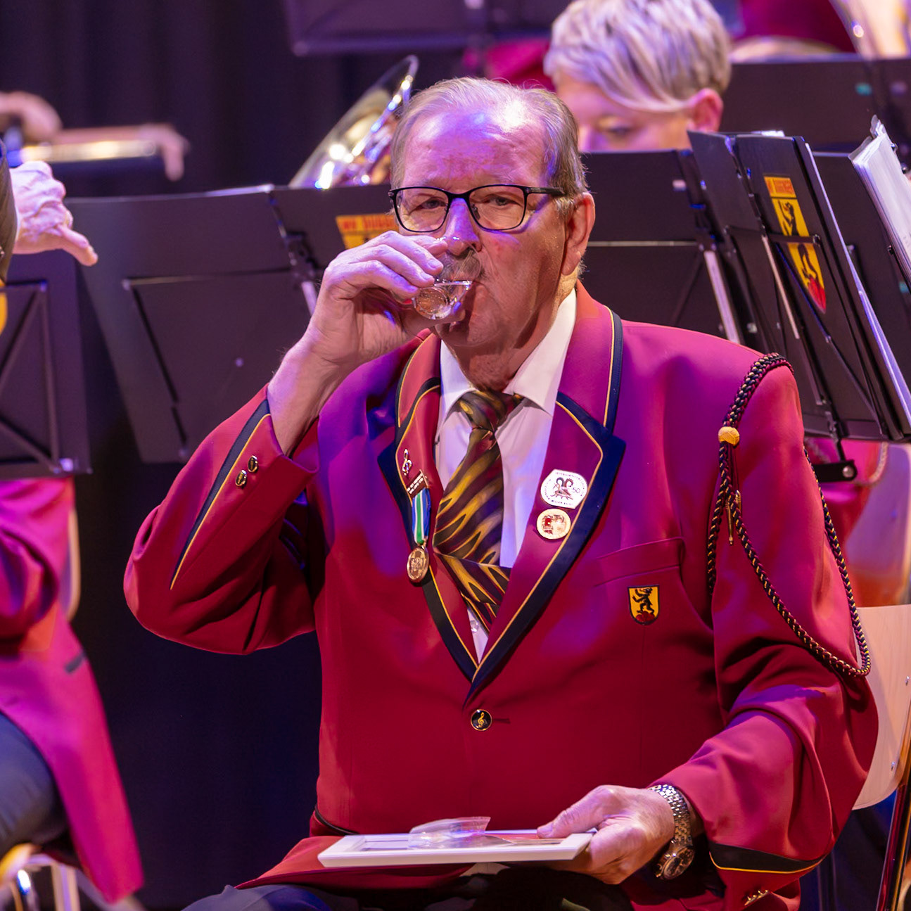 Herr [Name des Dirigenten/Musikers], ein Mitglied der Blaskapelle, nimmt einen Schluck aus einem kleinen Glas.  Er trägt die Uniform der Kapelle und hat mehrere Auszeichnungen an seiner Jacke.  Wahrscheinlich ist dies eine Pause während eines Konzerts oder einer Übung.
