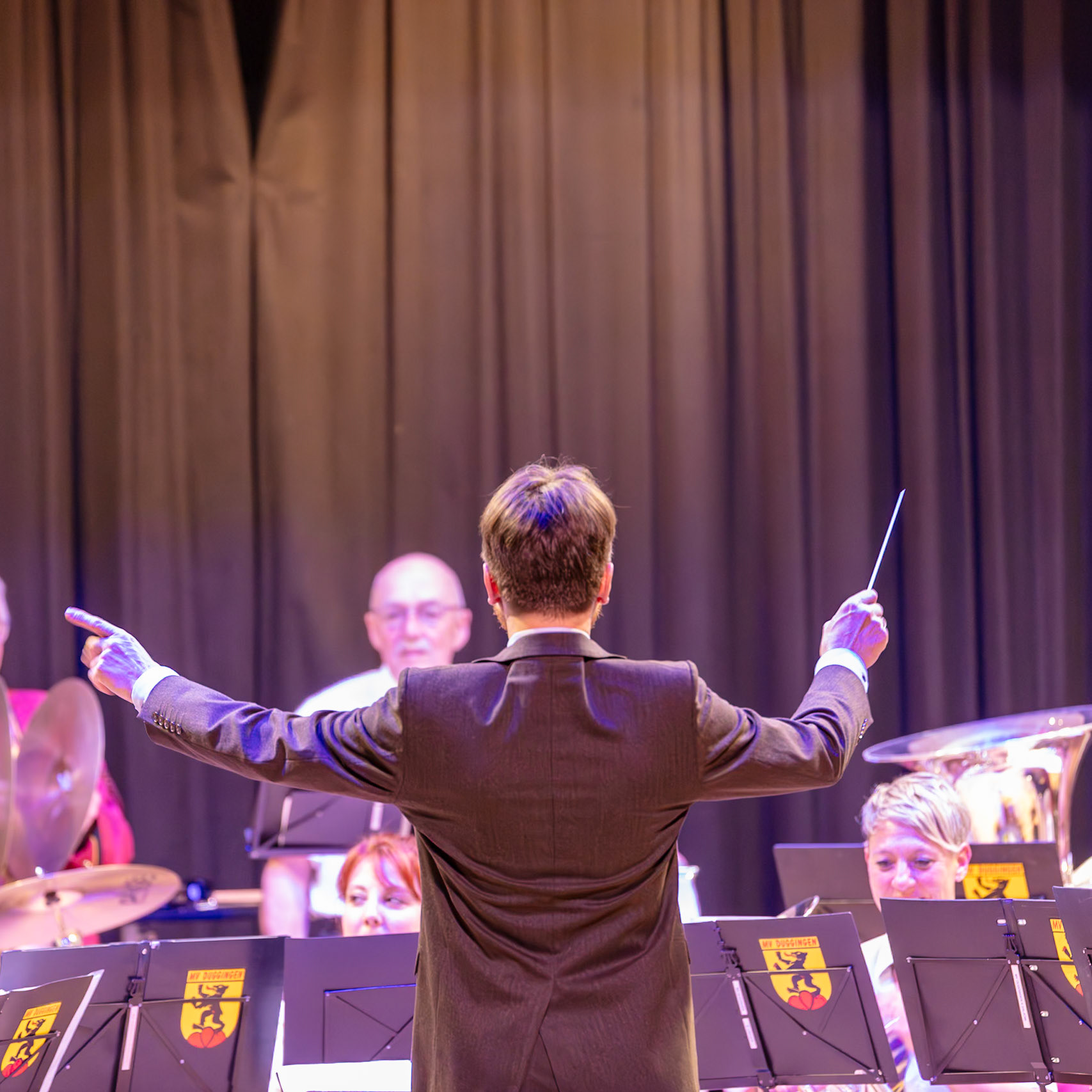 Ein Dirigent leitet ein Blasorchester. Herr [Name des Dirigenten, falls bekannt], der Dirigent, steht vor dem Blasorchester "MV Duggingen" und dirigiert mit einem Dirigentenstab.  Die Musiker des Orchesters spielen Musik, und der Dirigent gibt ihnen die Taktvorgabe.  Die Stimmung ist konzentriert und professionell.
