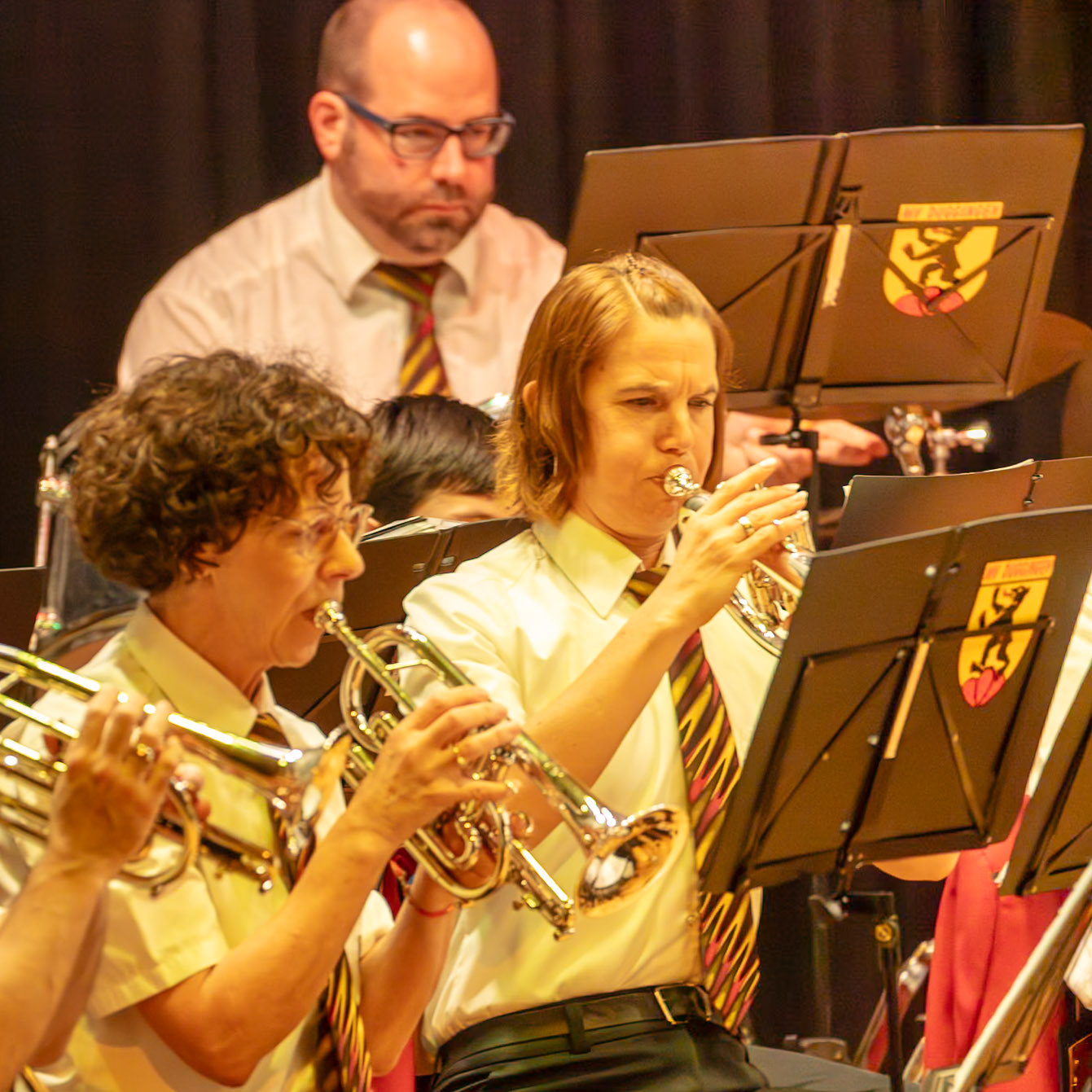Auf der Bühne spielen mehrere Musikerinnen und Musiker eines Blasorchesters.  Frau [Name der Frau, wenn bekannt], die eine Trompete spielt, und eine weitere Person, die eine Trompete spielt, sind gut zu erkennen.  Sie konzentrieren sich auf die Ausführung ihrer Musikstücke.  Das Orchester spielt vermutlich für ein Publikum, das nicht im Bild zu sehen ist.  Die Atmosphäre ist konzentriert und musikalisch.