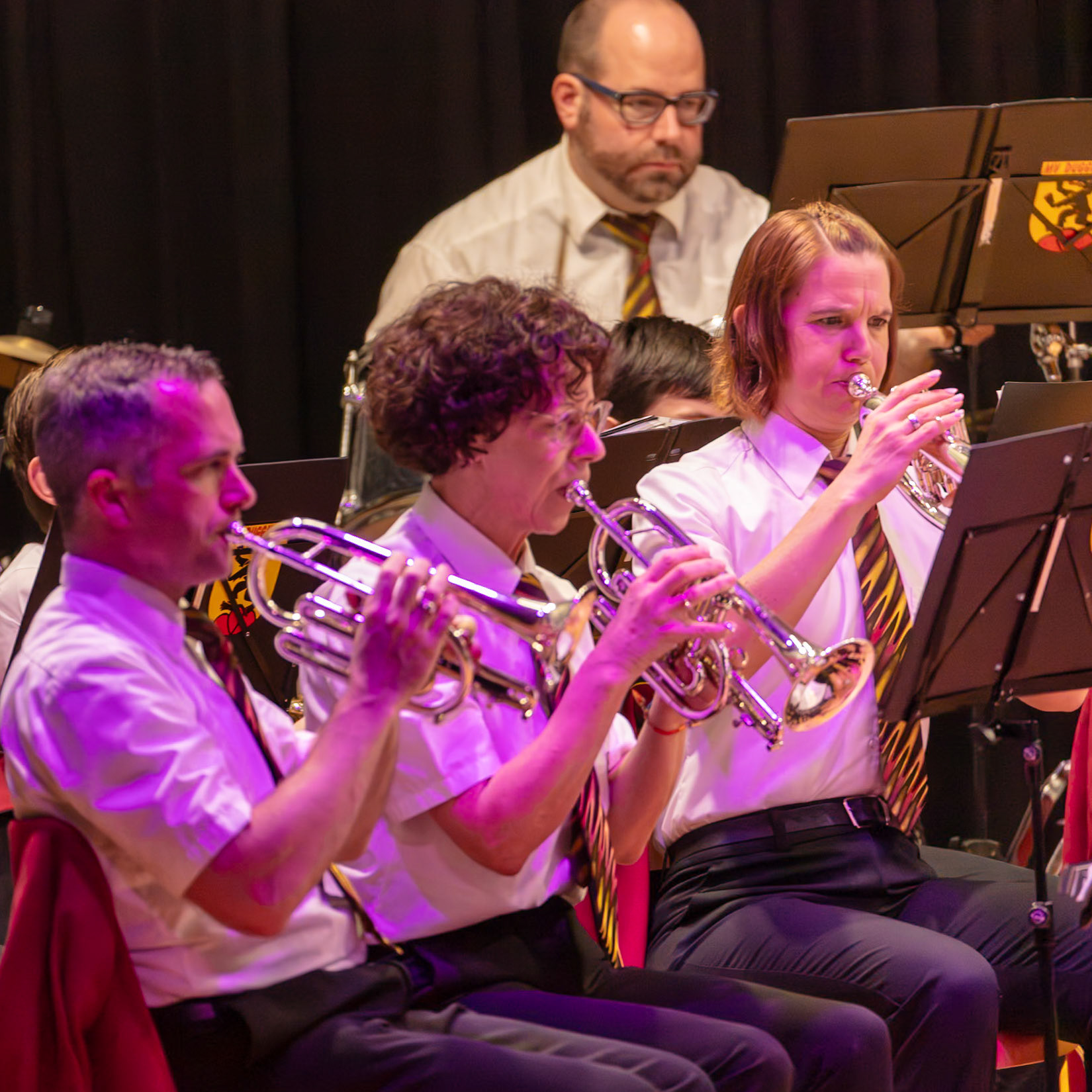 Eine Blaskapelle spielt auf der Bühne.  Frau [Name der Frau], die Trompete spielt, konzentriert sich auf die Musik.  Neben ihr spielt Herr [Name des Mannes], ebenfalls Trompete, und daneben spielt Frau [Name der Frau] ebenfalls Trompete.  Alle Musiker tragen einheitliche weiße Hemden und dunkle Hosen.  Die Atmosphäre ist konzentriert und musikalisch.