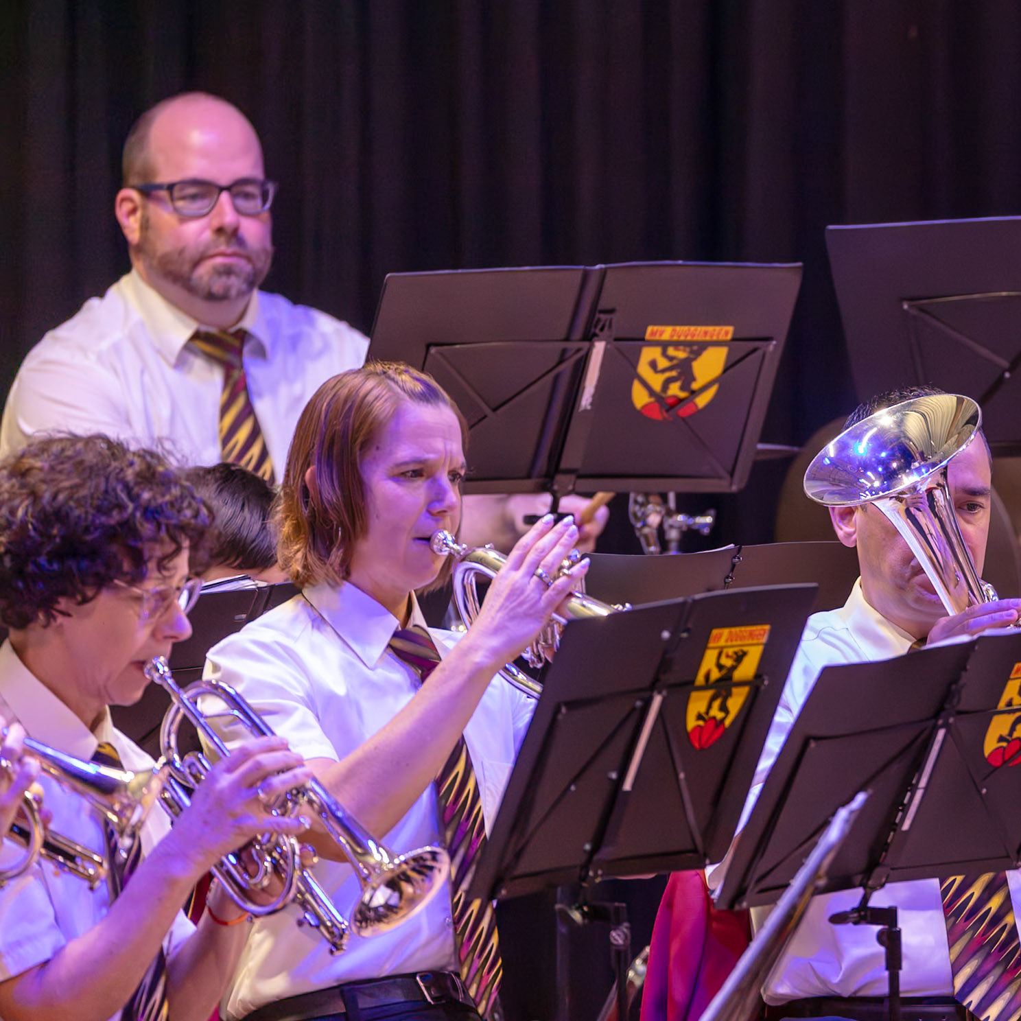 Ein Blasorchester spielt auf der Bühne.  Frau [Name der Frau], die Trompete spielt, und Herr [Name des Herrn], der Posaune spielt, sind  Mitglieder des Orchesters.  Sie alle tragen einheitliche, weiße Hemden und Krawatten.  Das Orchester spielt vermutlich eine Melodie, die von einem Dirigenten geleitet wird, der nicht im Bild zu sehen ist.  Die Musik wird in einem Konzertsaal oder einer ähnlichen Veranstaltung aufgeführt.