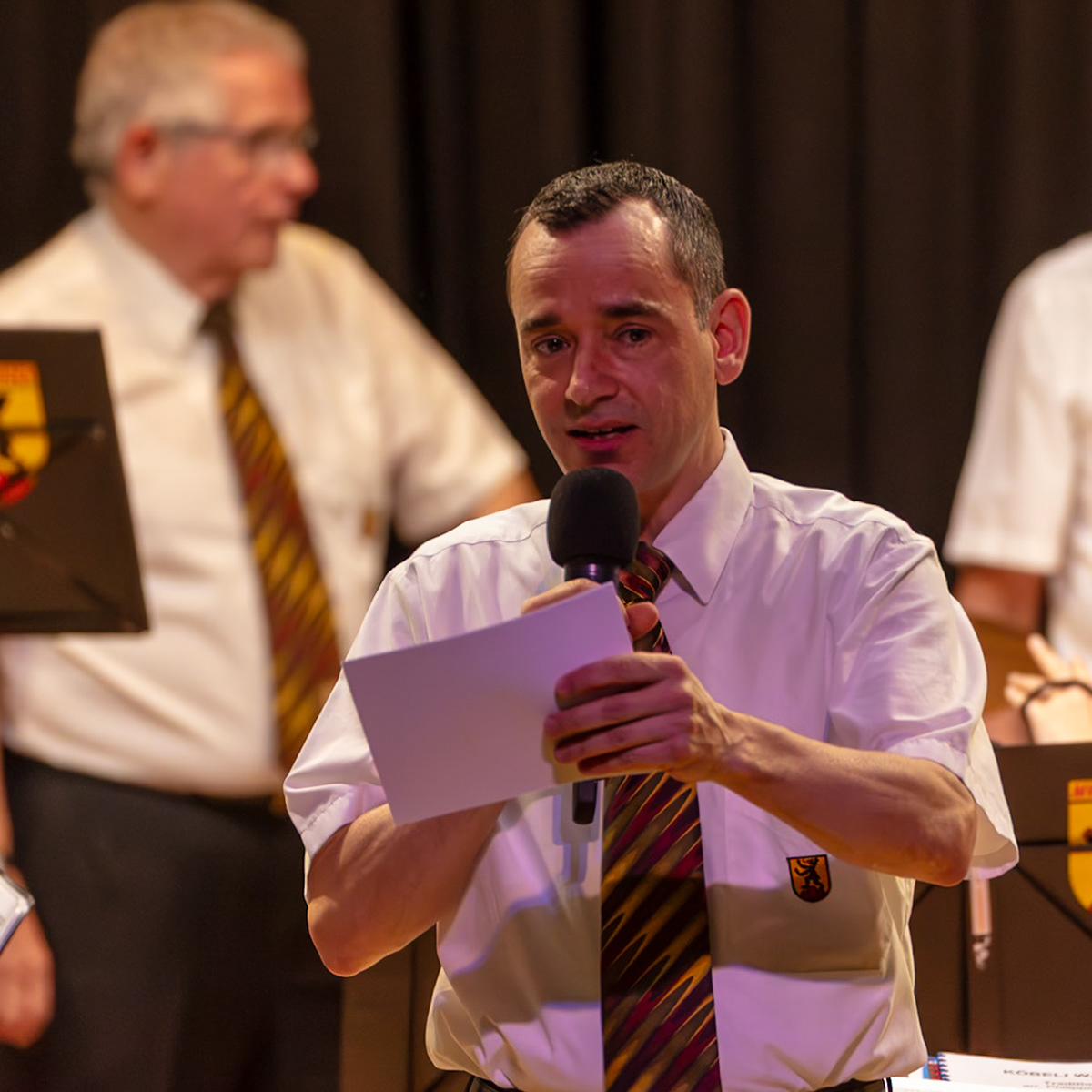 Herr [Name des Sprechers], vermutlich ein Mitglied des [Name des Vereins/der Organisation], hält eine Ansprache.  Er liest dabei von einem Zettel vor, der in seinen Händen liegt.  Der Anlass ist vermutlich eine Veranstaltung des Vereins, wie z.B. eine Mitgliederversammlung oder eine Feier.  Die Anwesenden scheinen dem Sprecher aufmerksam zuzuhören.