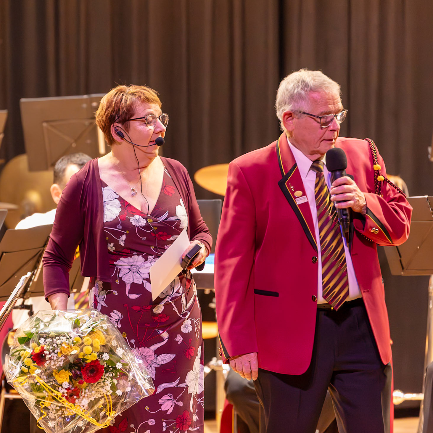 Auf der Bühne werden Personen geehrt. Frau [Name der Frau], vermutlich eine Dirigentin oder eine Person mit einer wichtigen Funktion im Orchester, überreicht Herrn [Name des Mannes], dem Dirigenten oder einem Ehrenmitglied des Orchesters, einen Blumenstrauß.  Die Geste symbolisiert Anerkennung und Wertschätzung für seine Leistungen.  Der Anlass könnte ein Konzert, ein Jubiläum oder eine ähnliche Feier sein.