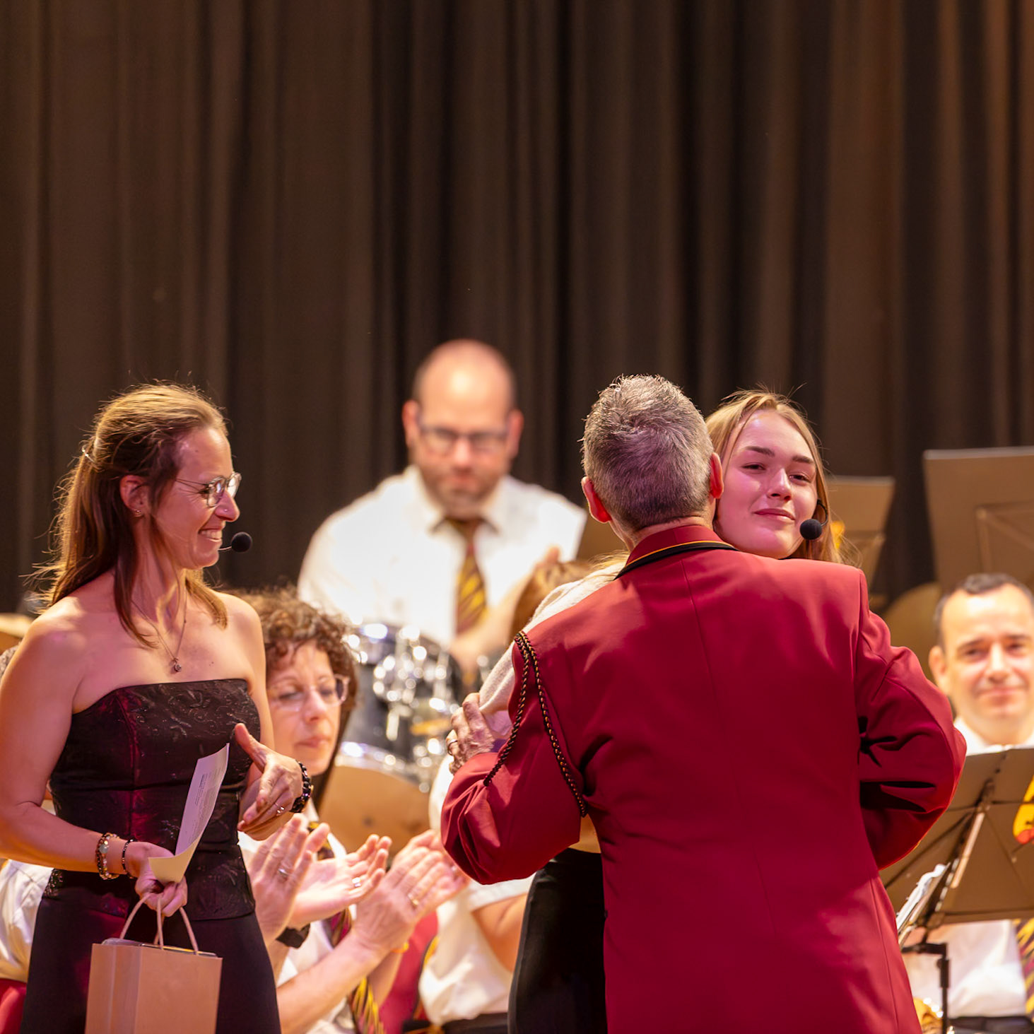 Eine junge Frau, vermutlich eine Musikerin, wird von einem Dirigenten oder einem anderen Musik-Funktionär herzlich umarmt.  Sie erhält eine Auszeichnung oder ein Geschenk, wie die braune Geschenkverpackung und die applaudierenden Hände andeuten.  Die Geste symbolisiert die Anerkennung und Wertschätzung ihrer Leistung oder ihres Engagements im Rahmen eines Musik- oder Orchester-Events.