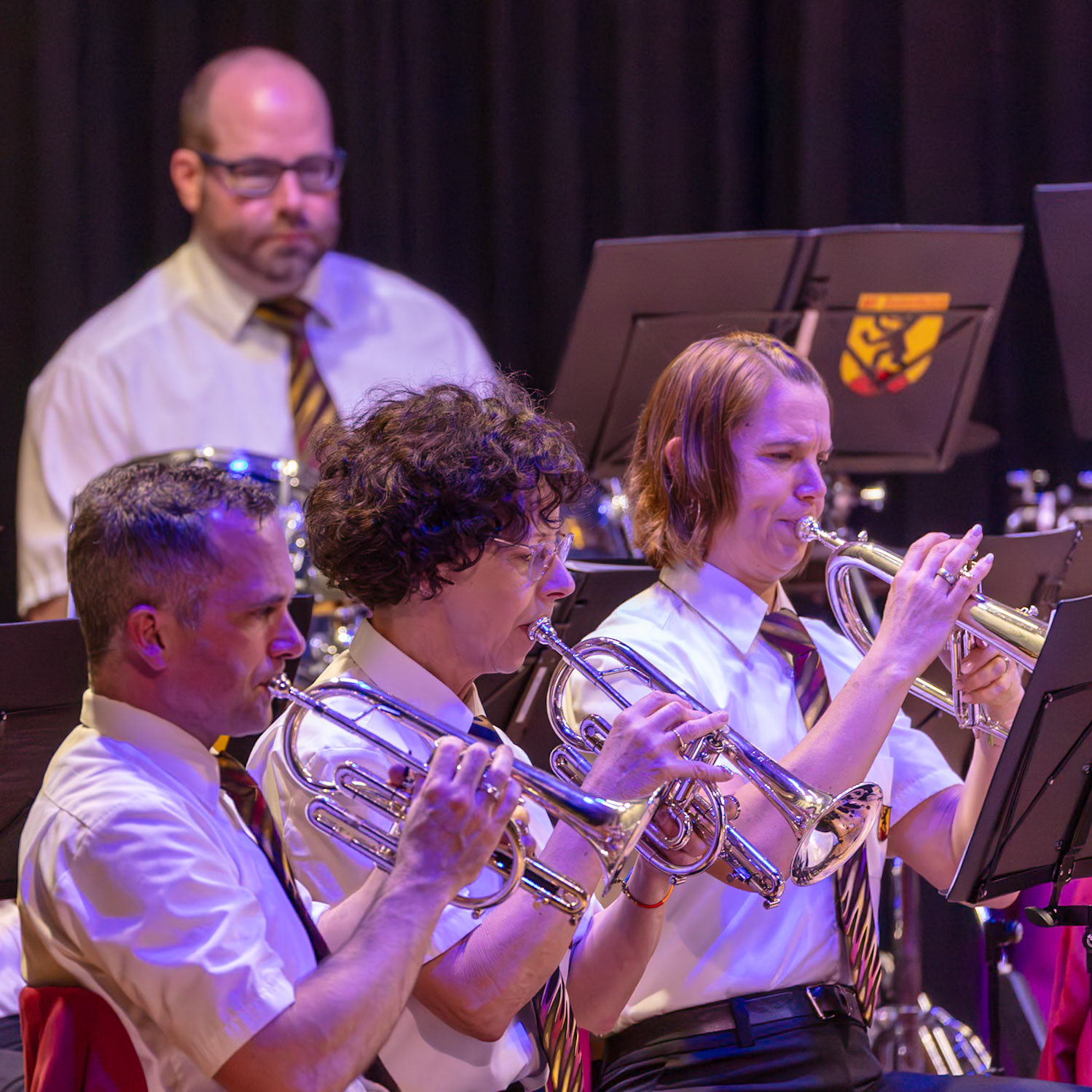 Eine Blaskapelle spielt.  Mehrere Musikerinnen und Musiker, darunter [Name des Mannes 1] (Trompeter), und [Name der Frau 2] (Trompeterin), spielen auf ihren Instrumenten.  Sie tragen einheitliche weiße Hemden und Krawatten.  Die Musik wird in einem Konzertsaal oder einer ähnlichen Veranstaltung aufgeführt.  Der Fokus liegt auf dem gemeinsamen Musizieren und der musikalischen Leistung.