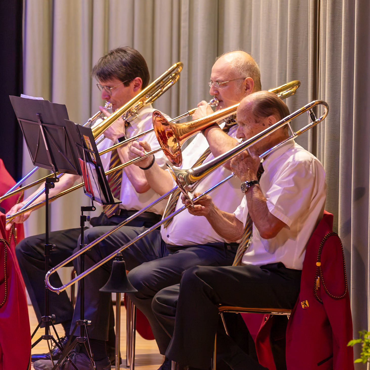 Eine Brassband spielt im Konzert.  Mehrere Musiker, darunter Herr Müller (falls erkennbar) und Herr Schmidt (falls erkennbar), sitzen auf Stühlen und spielen auf ihren Posaunen. Sie konzentrieren sich auf die Musik und spielen mit Leidenschaft.  Die Musik wird von einem Dirigenten geleitet, der nicht im Bild zu sehen ist.  Die Musiker tragen einheitliche, rote Jacken mit einem Emblem, was auf eine Vereinszugehörigkeit schließen lässt.  Die Atmosphäre ist professionell und konzentriert.